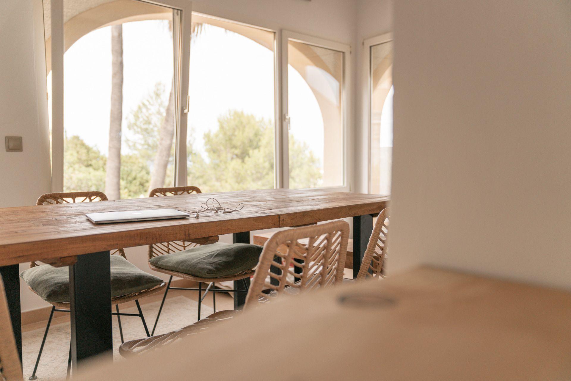 Large indoor coworking table with laptop and chairs, arched windows overlooking garden with palm trees