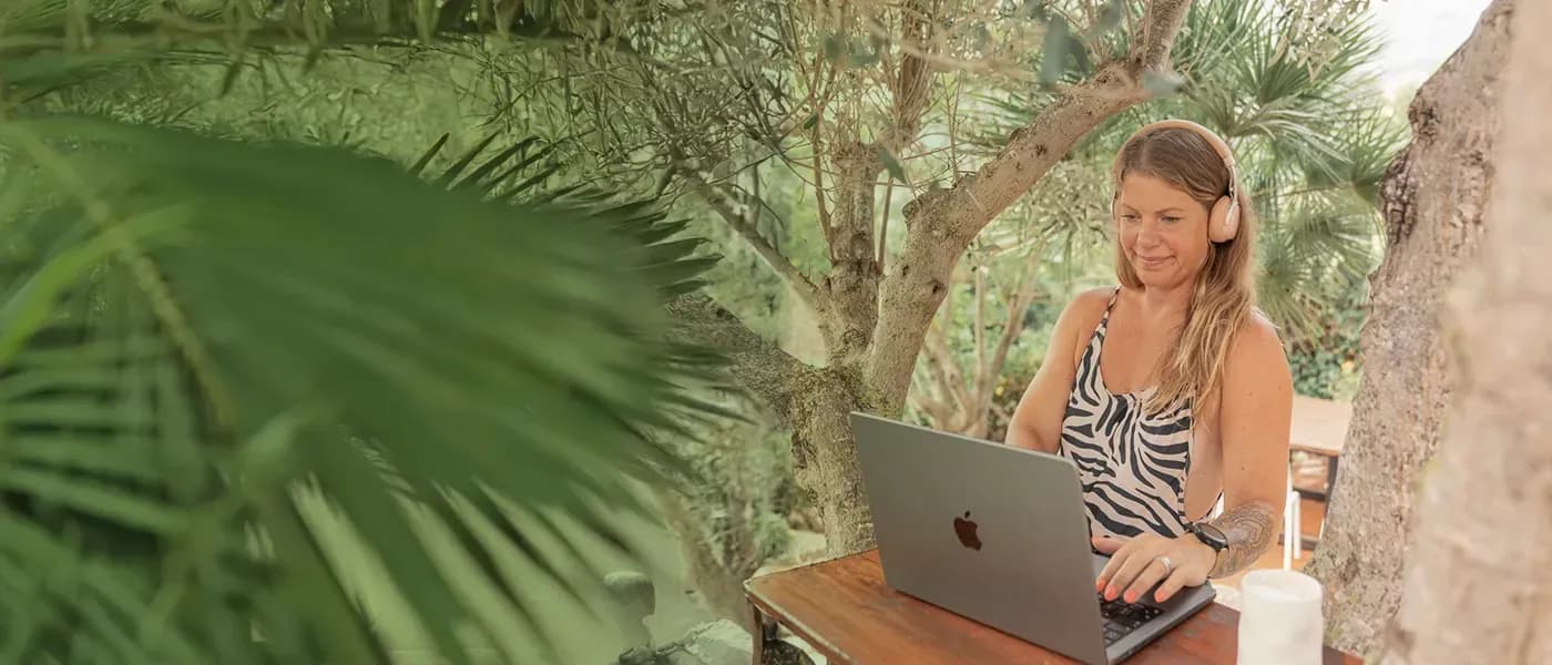 Person working on a laptop with headphones at the outdoor tree desk in the garden, surrounded by palm trees and Mediterranean vegetation