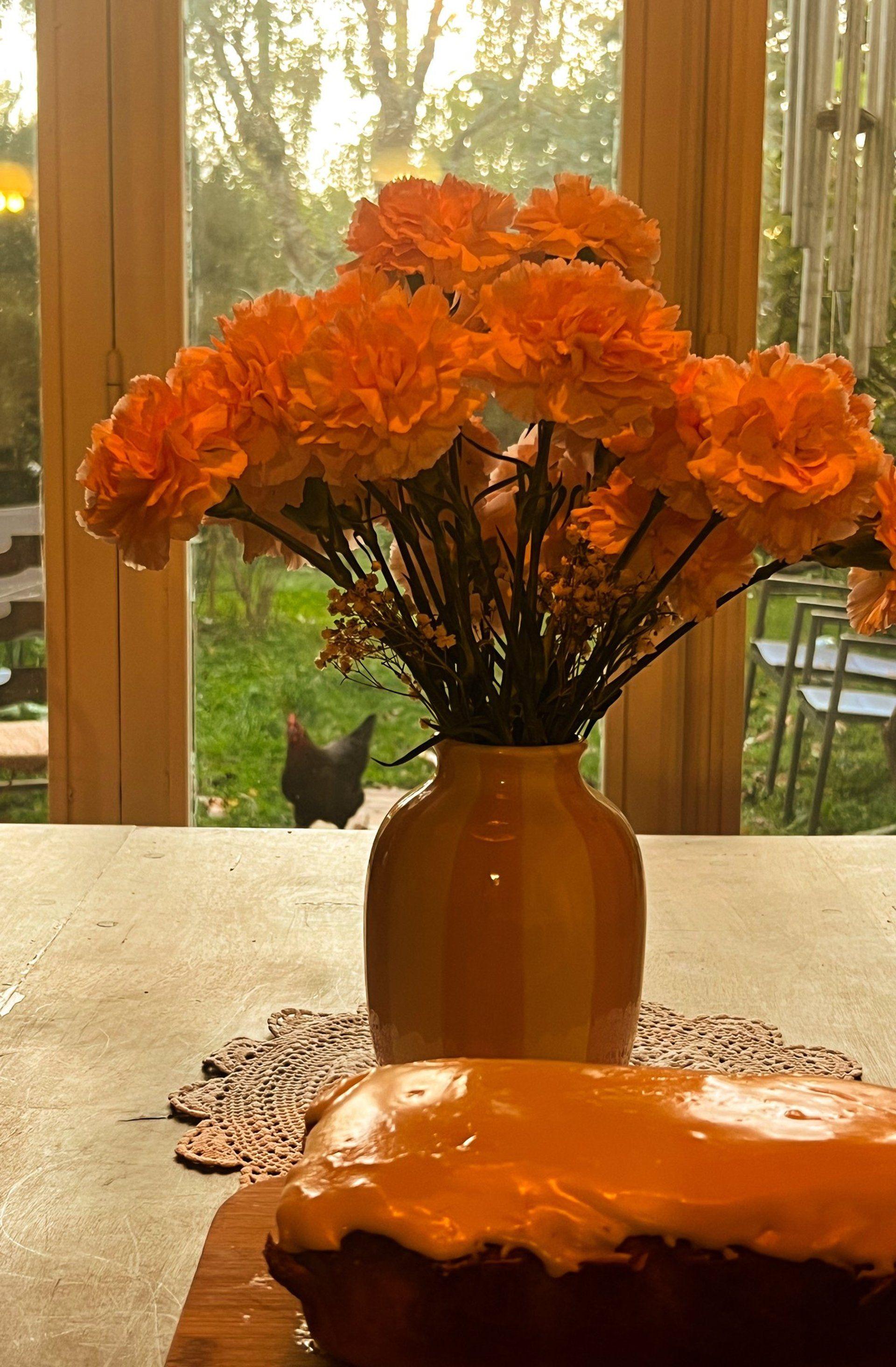 A kitchen table with a vase of orange flowers and a glazed cake, with a chicken visible through the window and the garden beyond