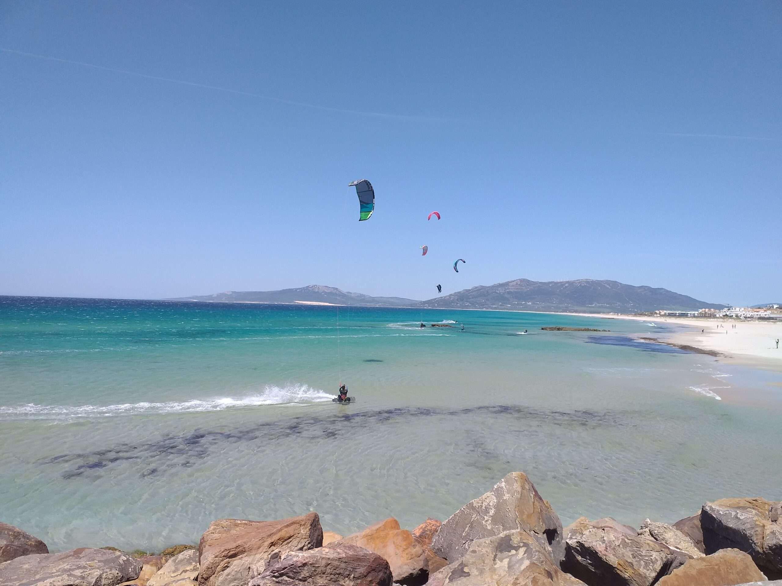 Rocks, crystal blue water, white sandy beach, 5 people kitesurfing, mountains in the back