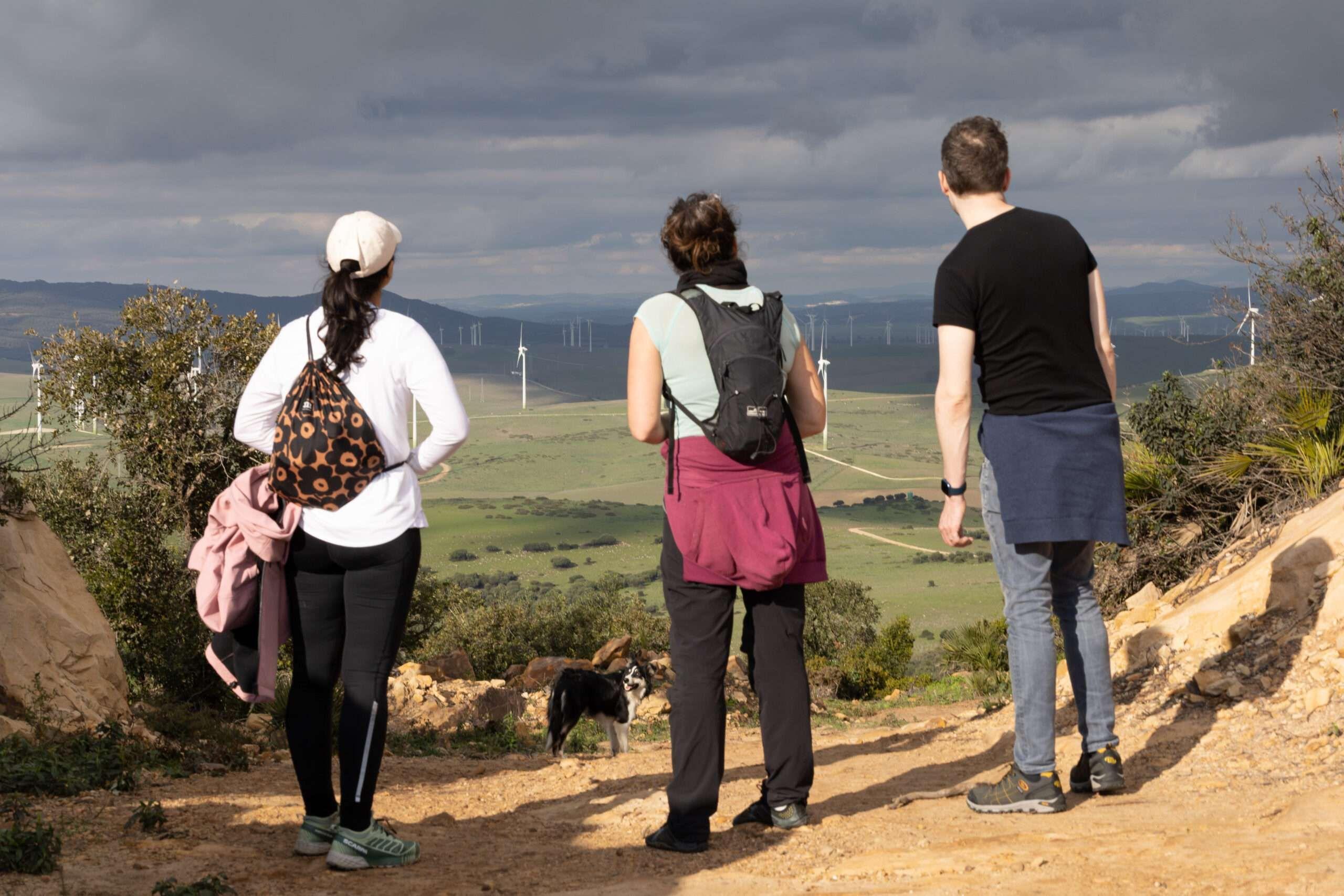 3 people standing on a gravel path with the back to us, looking over the green fields and hills with windmills below, a dog is standing a bit further down the path.