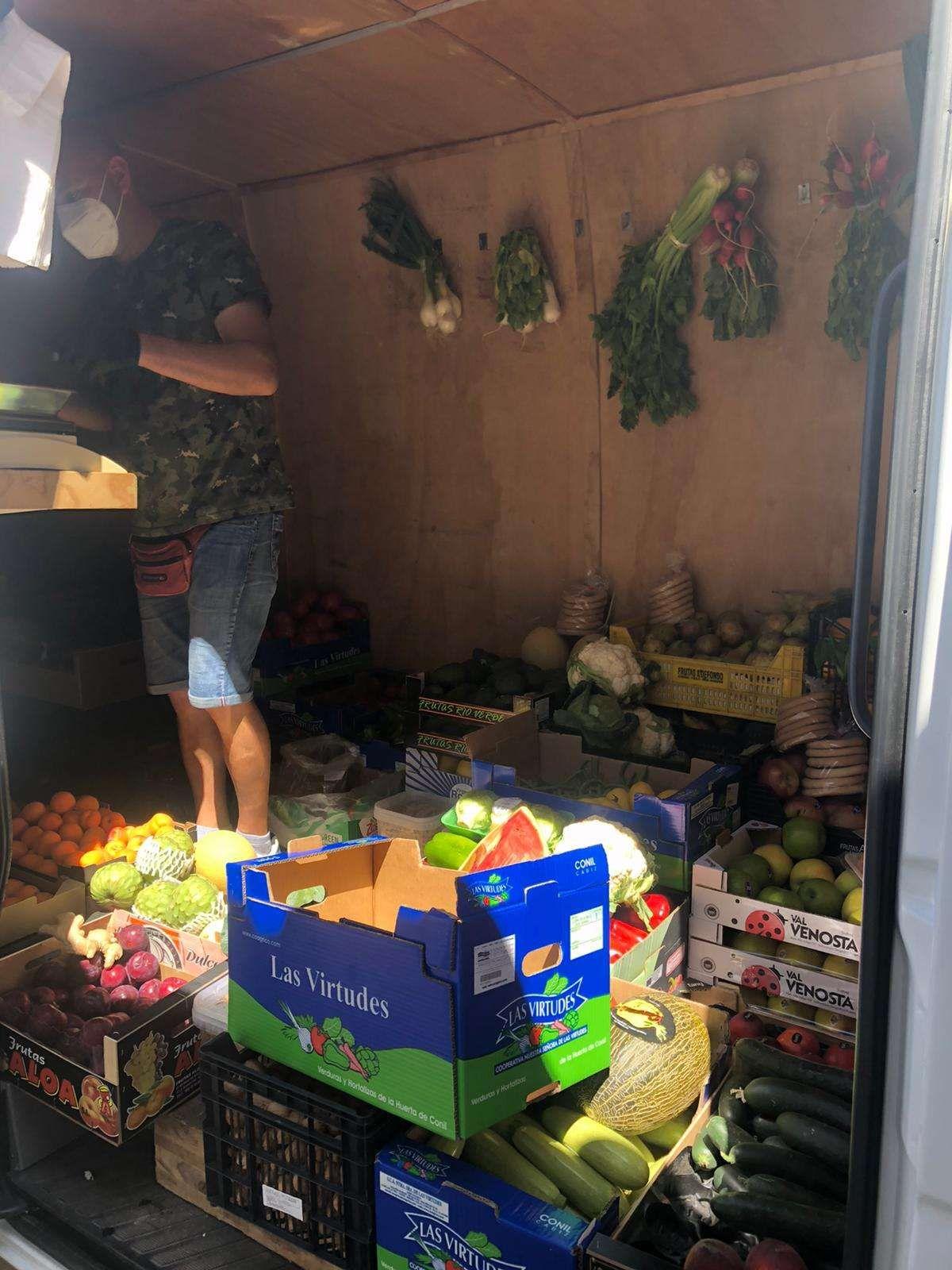 a big van with the side door wide open exposing plenty of boxes and cartons with all different kind of fruit and vegetable. A man standing in it with a scale.