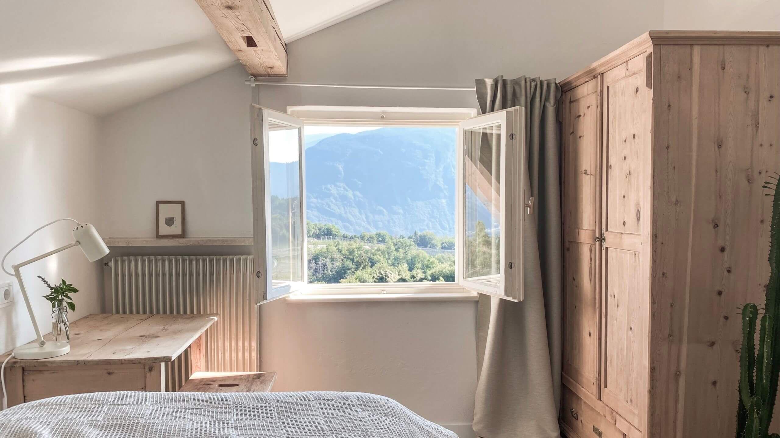 Cozy single bedroom with wooden desk, wardrobe, open window with mountain view, and exposed wooden beam ceiling