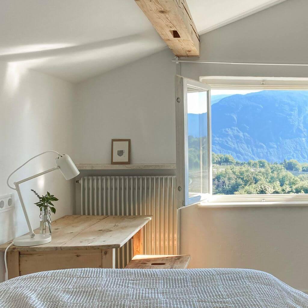 Bright bedroom with wooden desk, open window revealing mountain views, and exposed beam ceiling