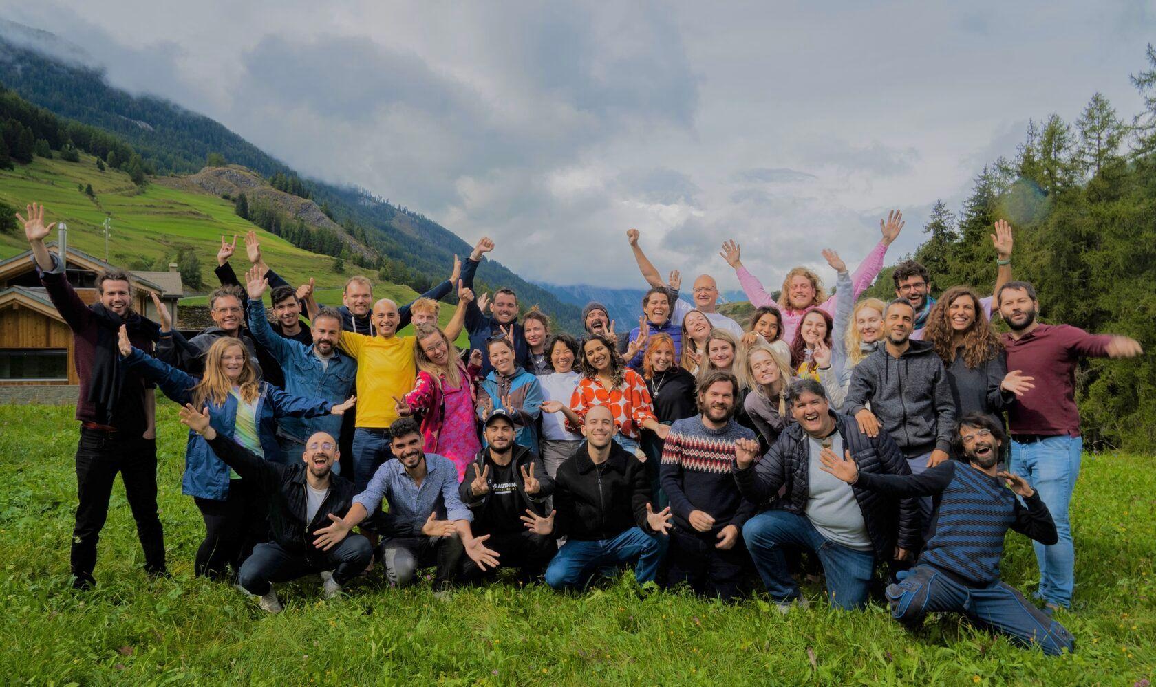 Large group of coliving community members posing together on a green alpine meadow with mountains and forest in the background