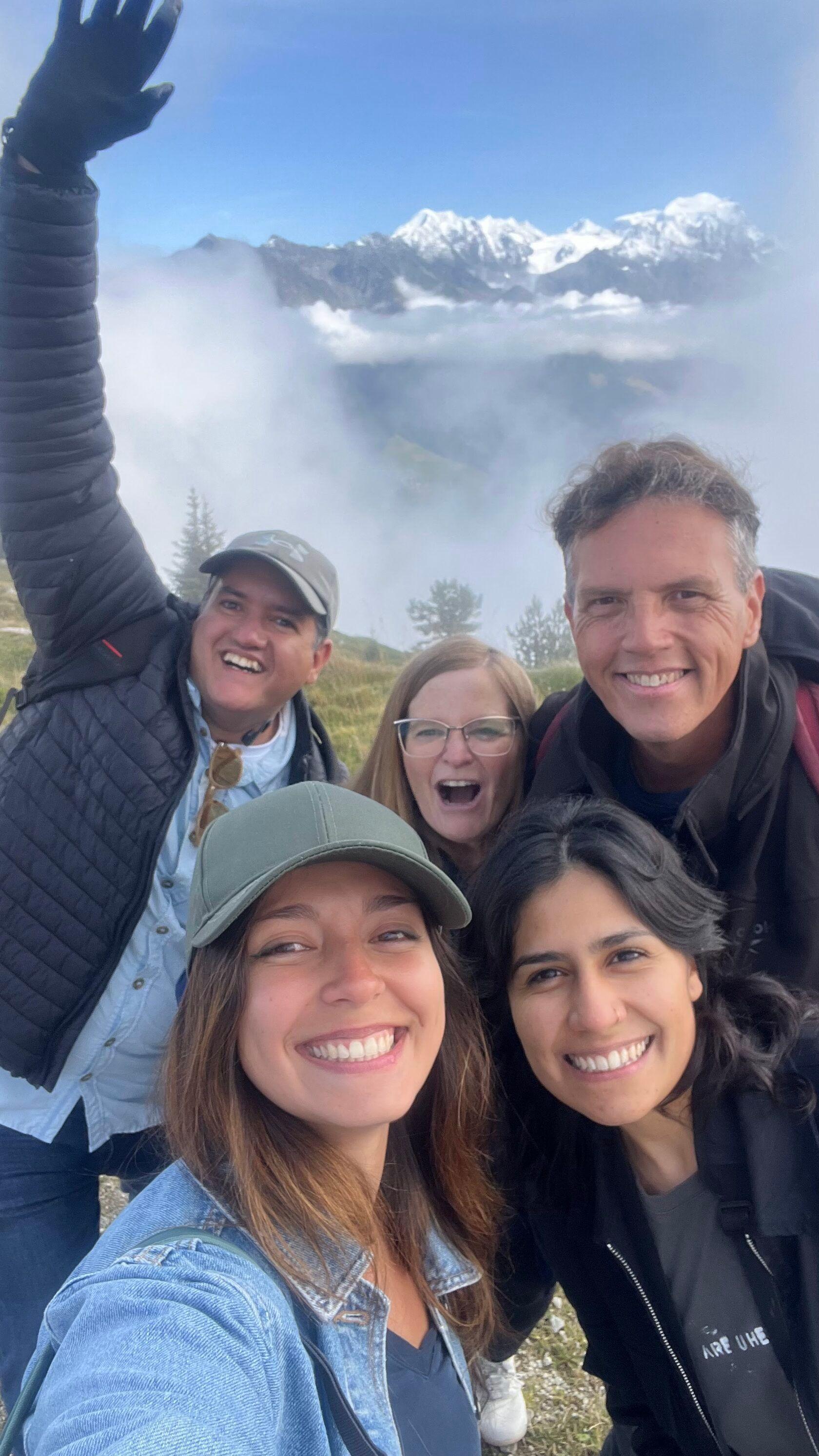 Group of five community members taking a selfie on a mountain hike with snowy peaks and clouds in the background