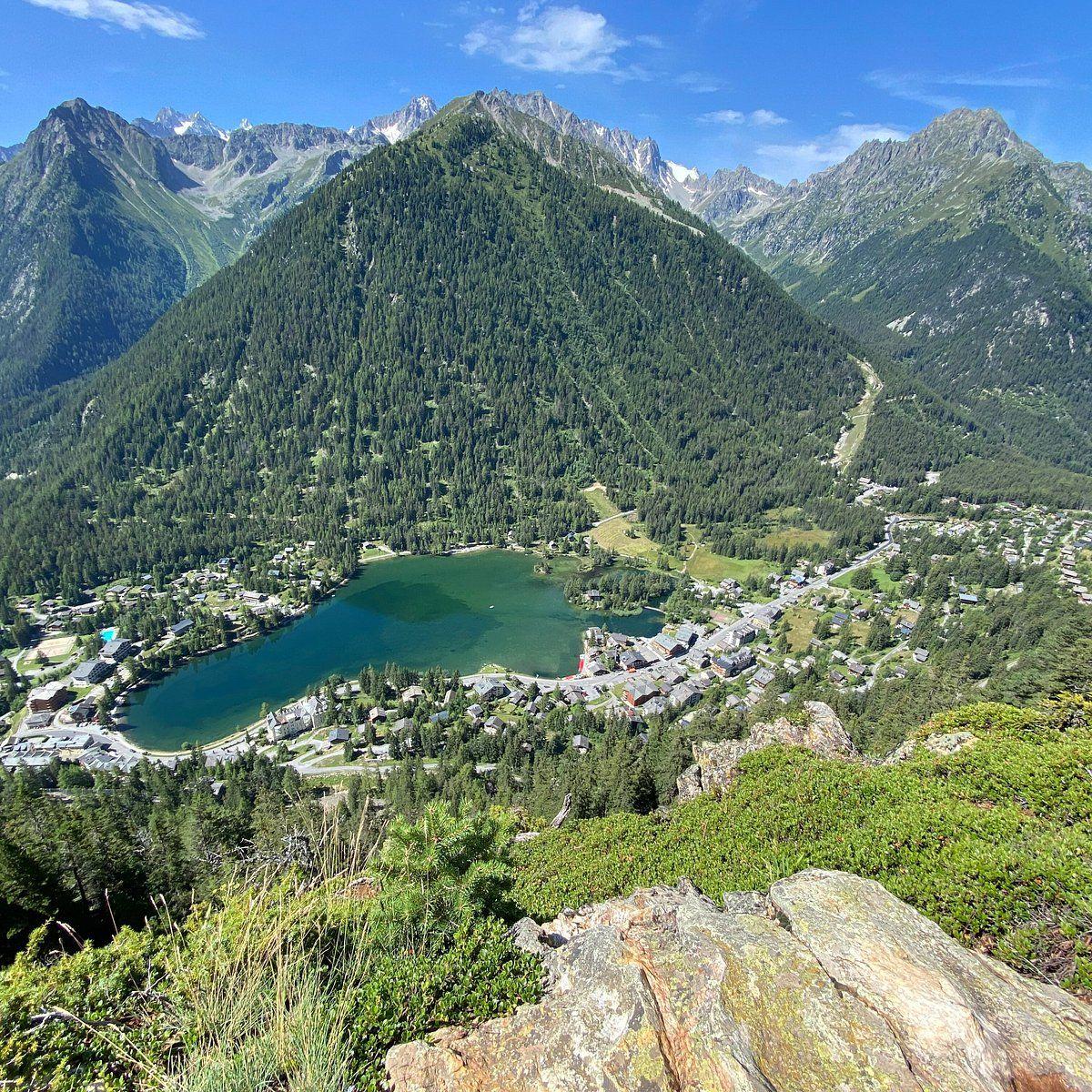 Aerial view of an alpine lake surrounded by mountains and a village, taken from a high vantage point during summer