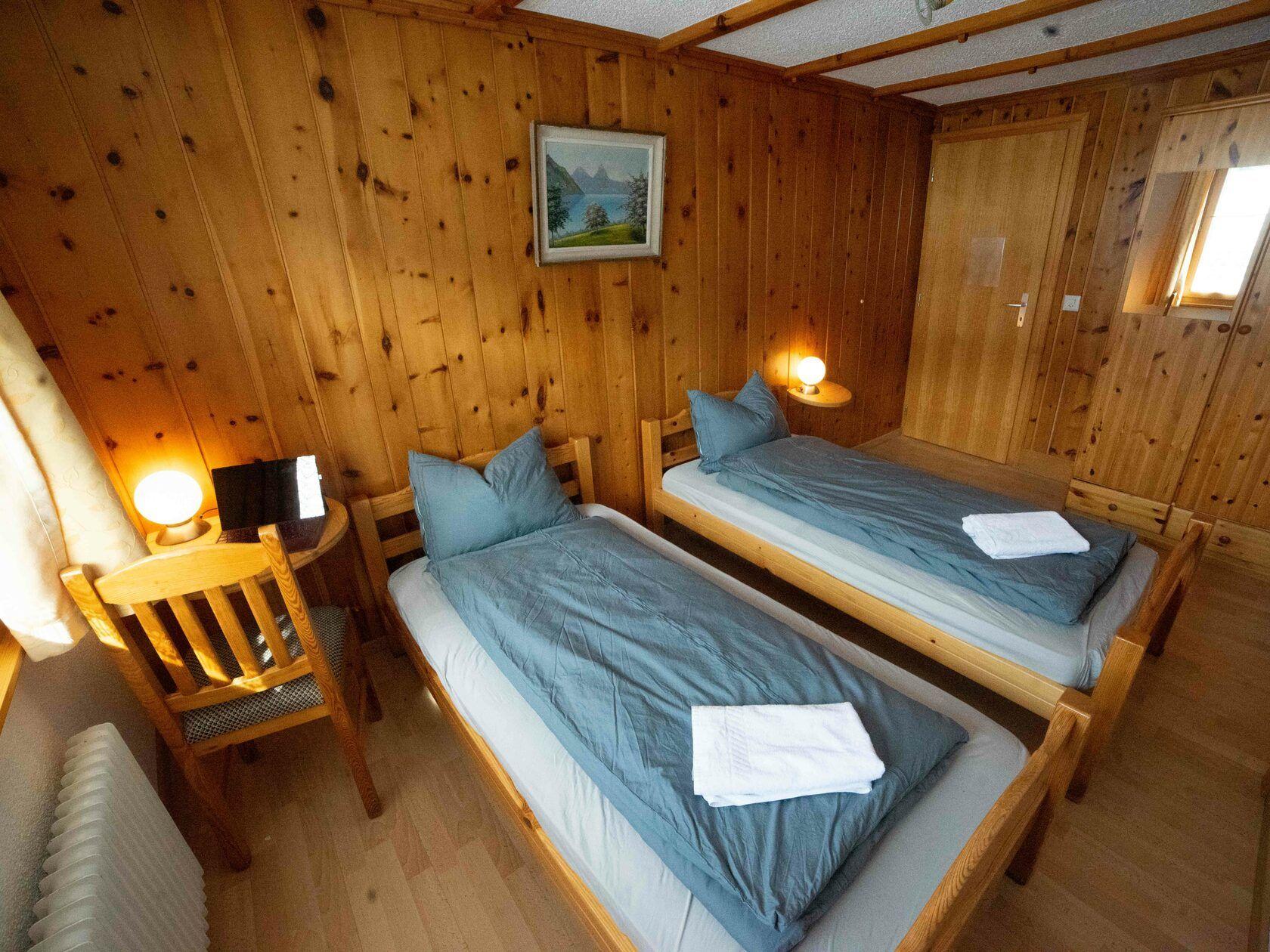 Cozy private room with two twin beds, wooden paneled walls, warm lighting, and a small desk