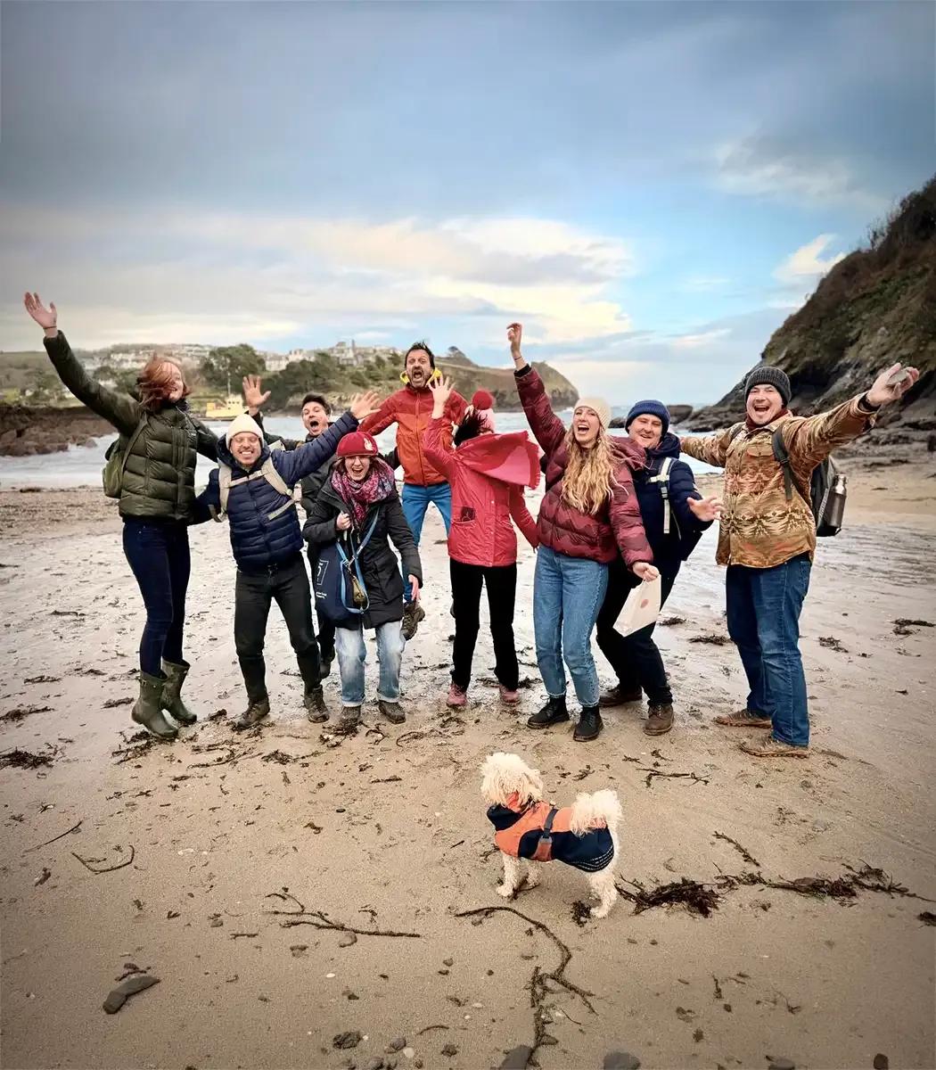 Group of Froomies residents on a beach outing in Cornwall, cheering with arms raised and a dog in front