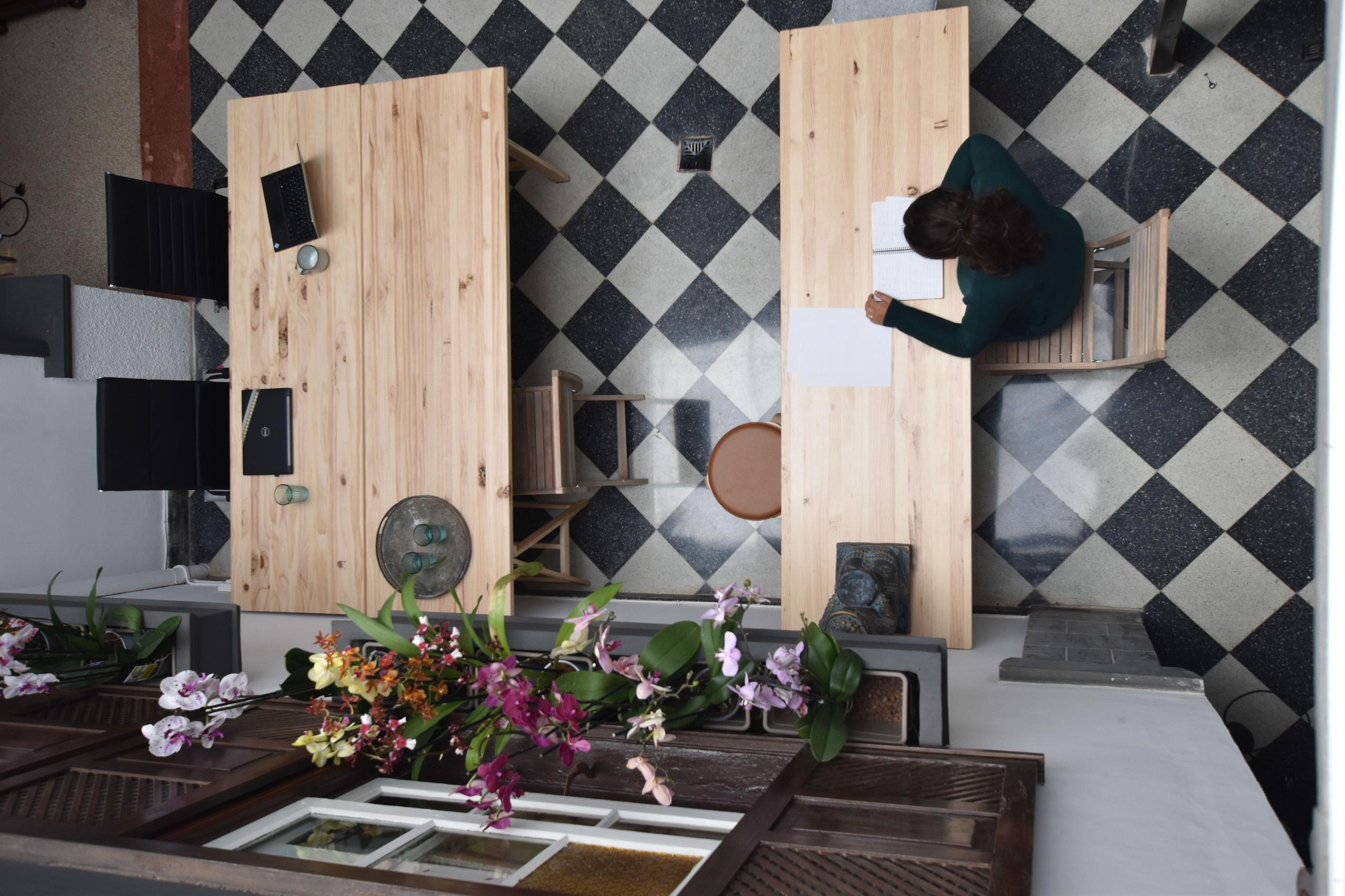 Overhead view of the communal coworking area with wooden desks, checkered tile floor, orchids, and a person working