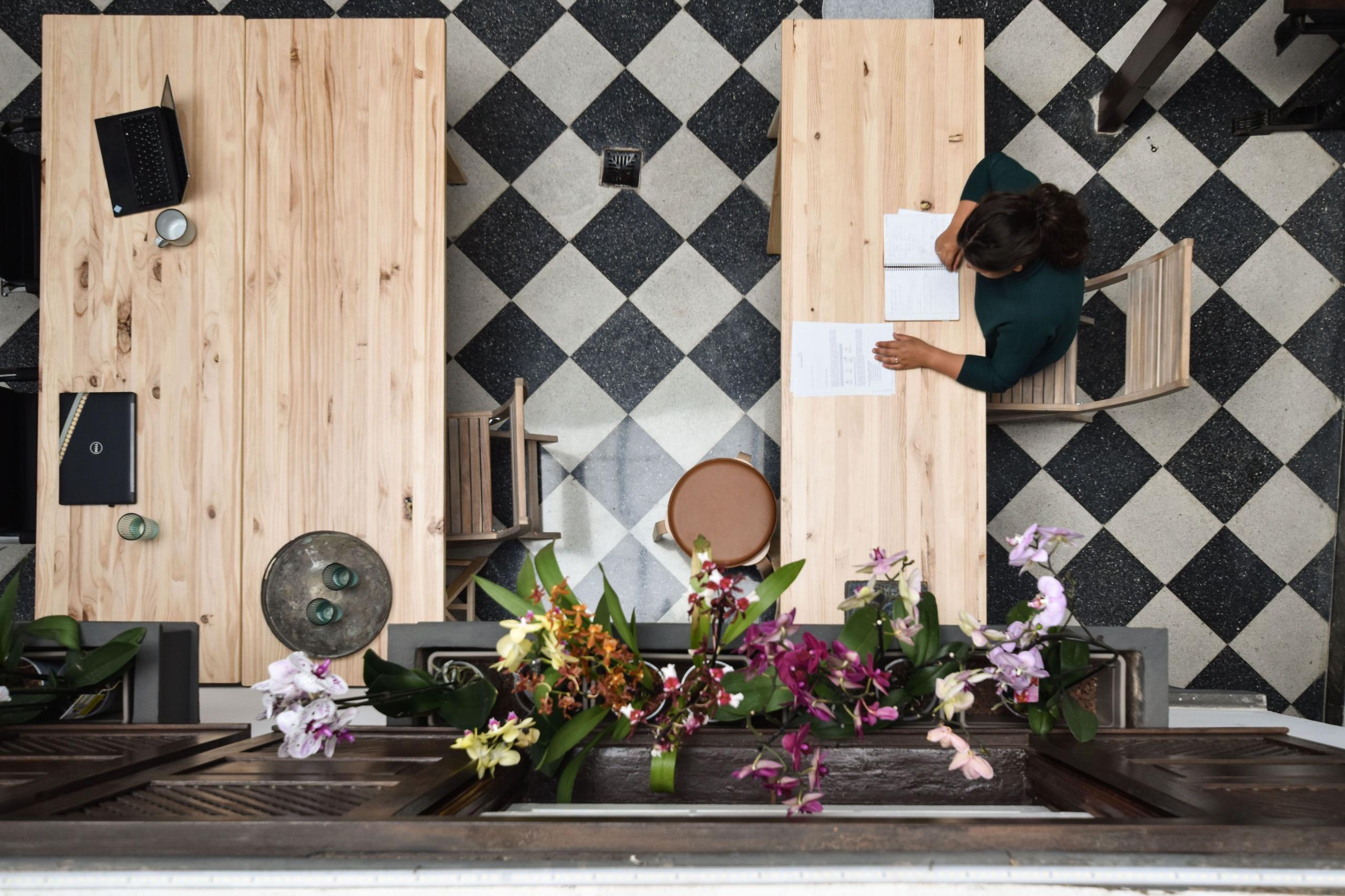 Aerial view of the communal coworking area showing wooden desks, checkered tile floor, and a person working, with orchid flowers in the foreground