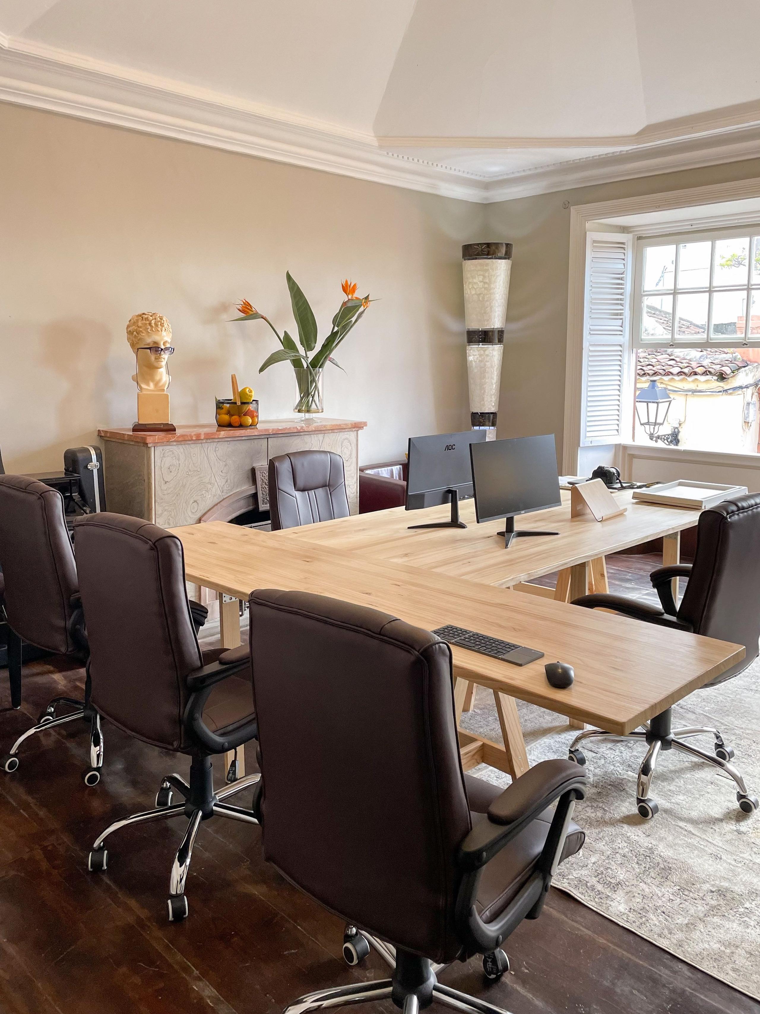 Spacious coworking room with large wooden desk, ergonomic office chairs, monitor, and large window with shutters overlooking the street