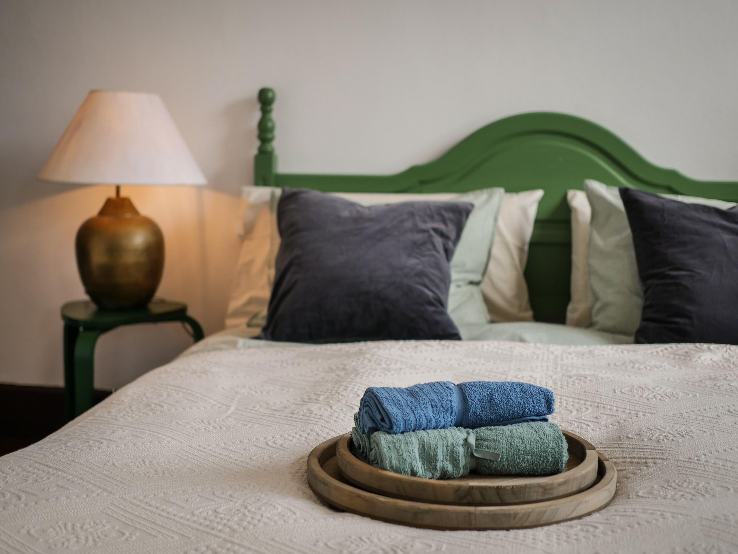 Double bed with green painted headboard, grey and mint pillows, folded towels on a wooden tray, and brass lamps on matching green side tables