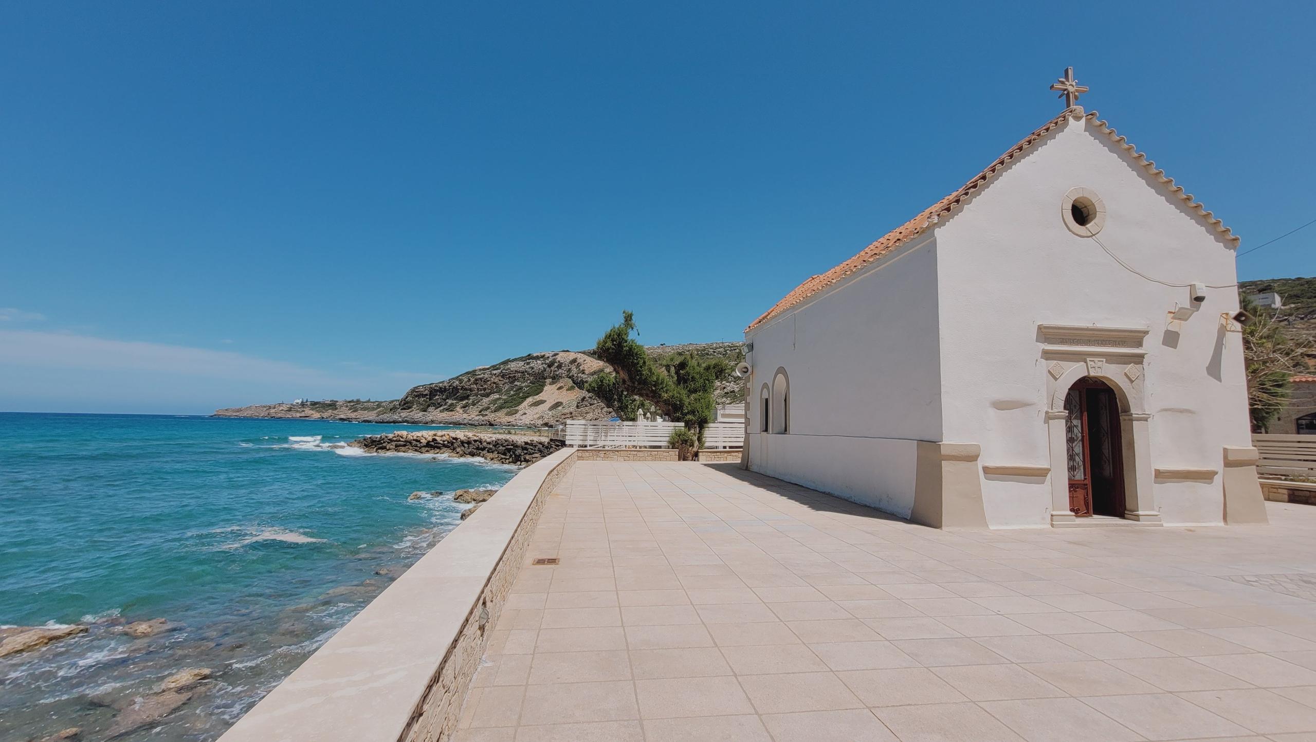 White chapel with a red roof by the sea, bright blue sky, and turquoise water. Stone pathway leads to greenery-covered hills in the background.