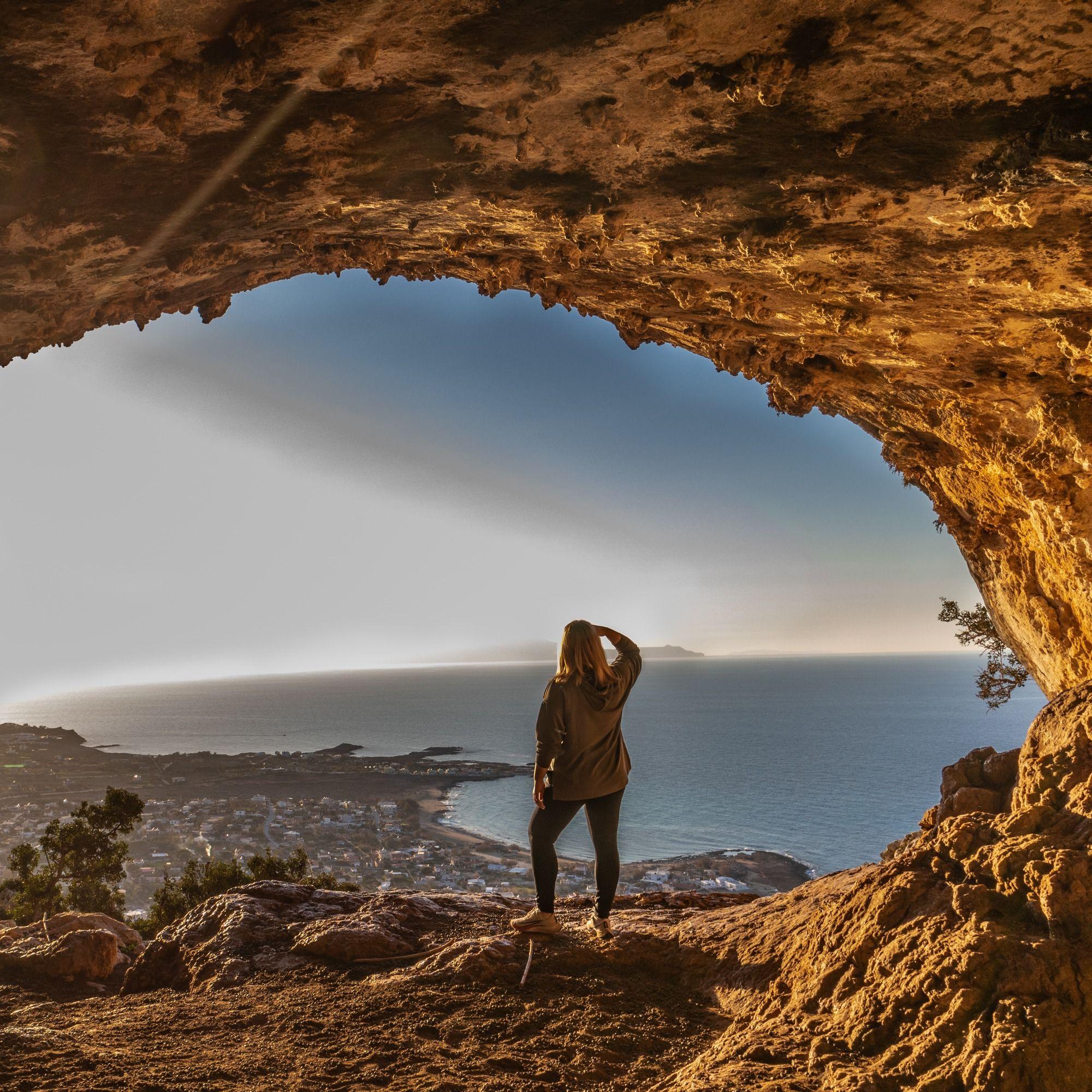 Person stands in a rocky cave entrance, overlooking a coastal town and ocean.