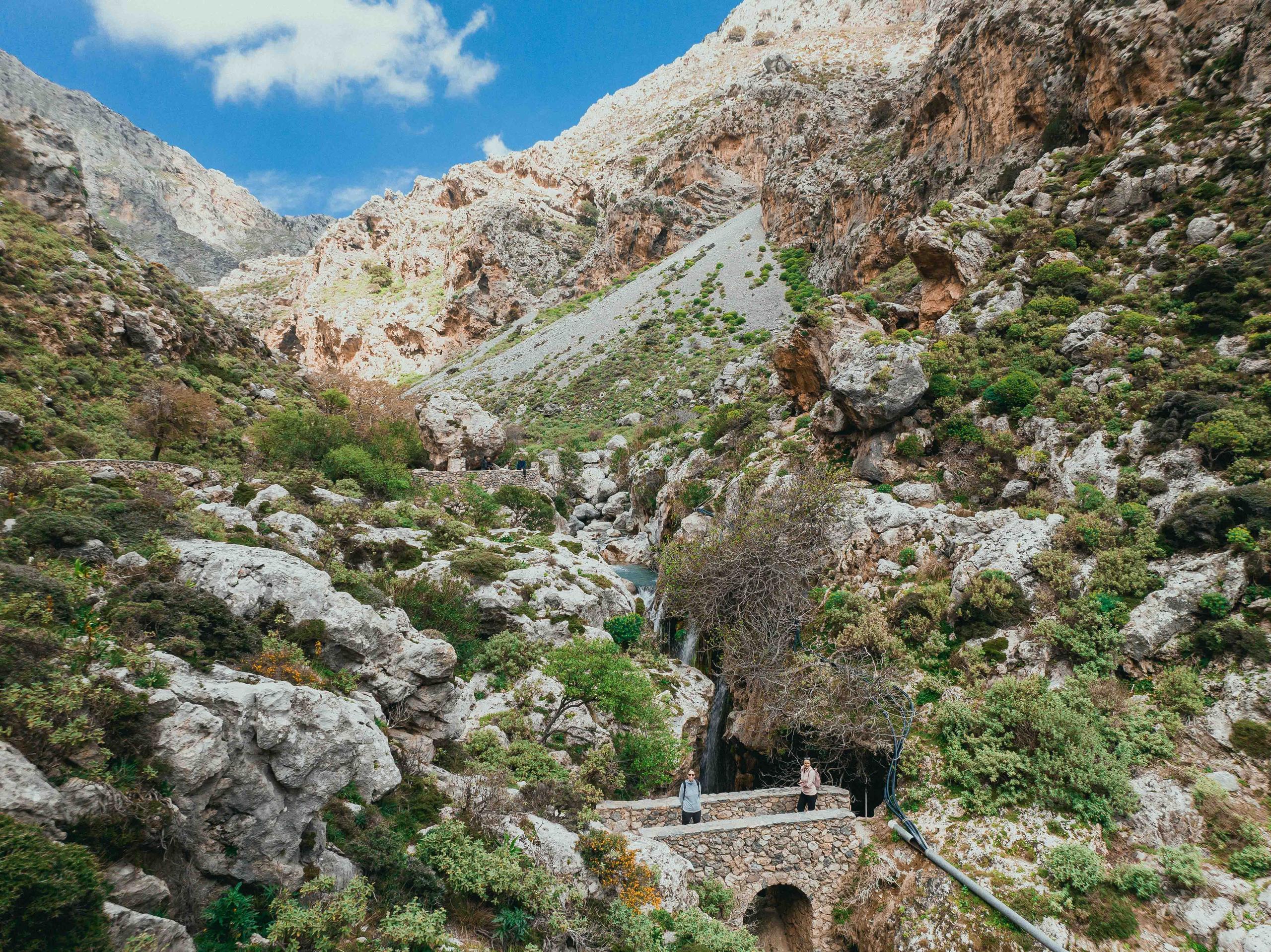 Rocky mountain landscape with green foliage, a stone bridge, and two people standing.