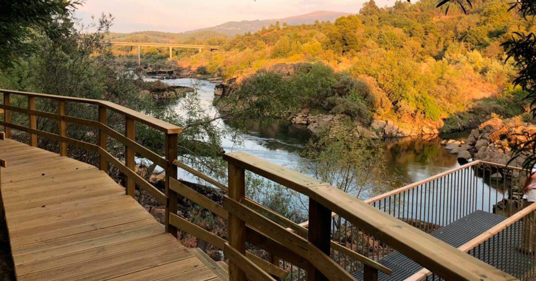 Wooden boardwalk along the Miño river with autumn foliage and a bridge in the background