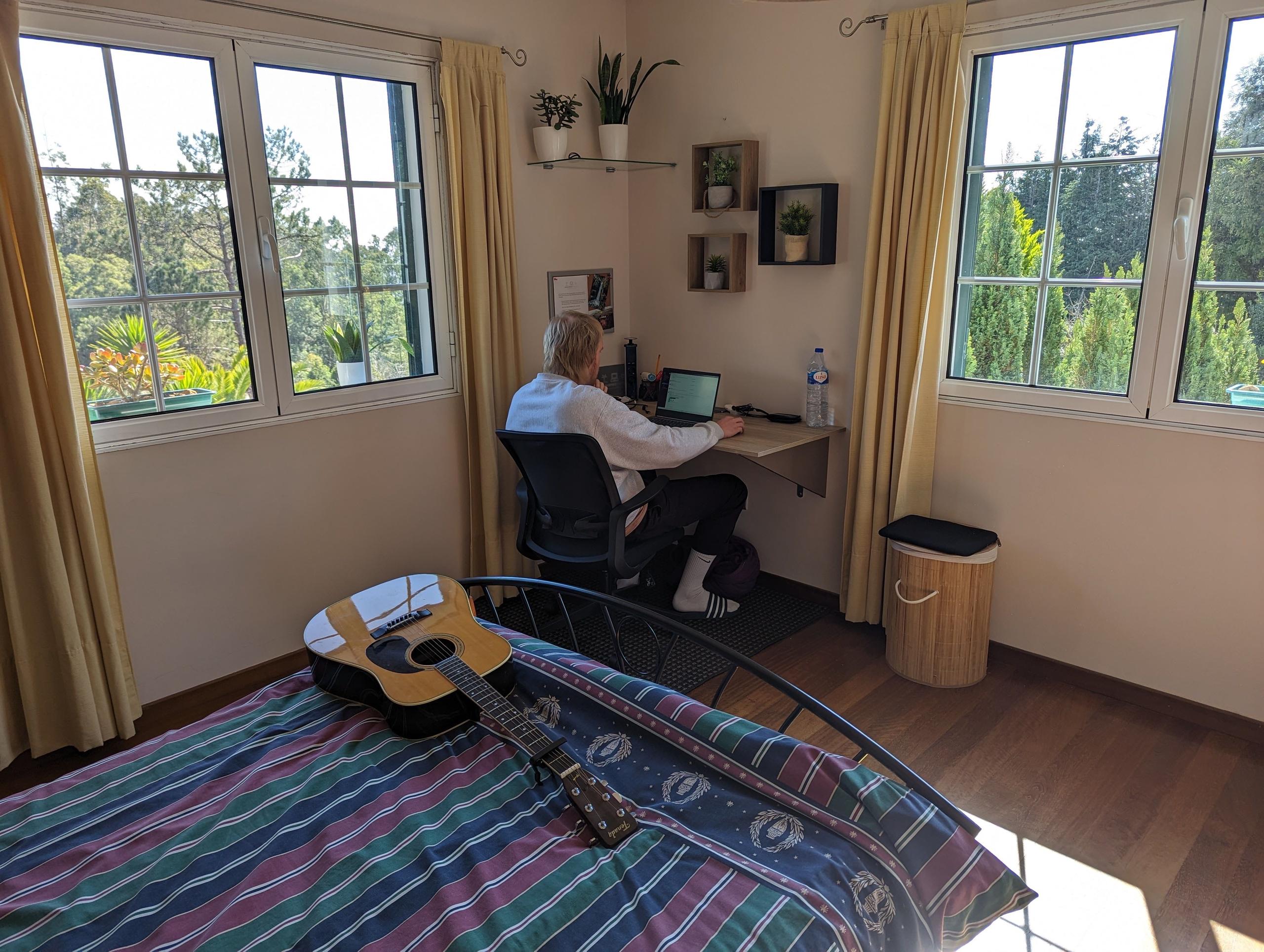 Enjoy a serene work environment like Paul, as he works in a beautiful double room with stunning south and east views, while his guitar rests on the bed, ready for a creative break at home office Madeira.