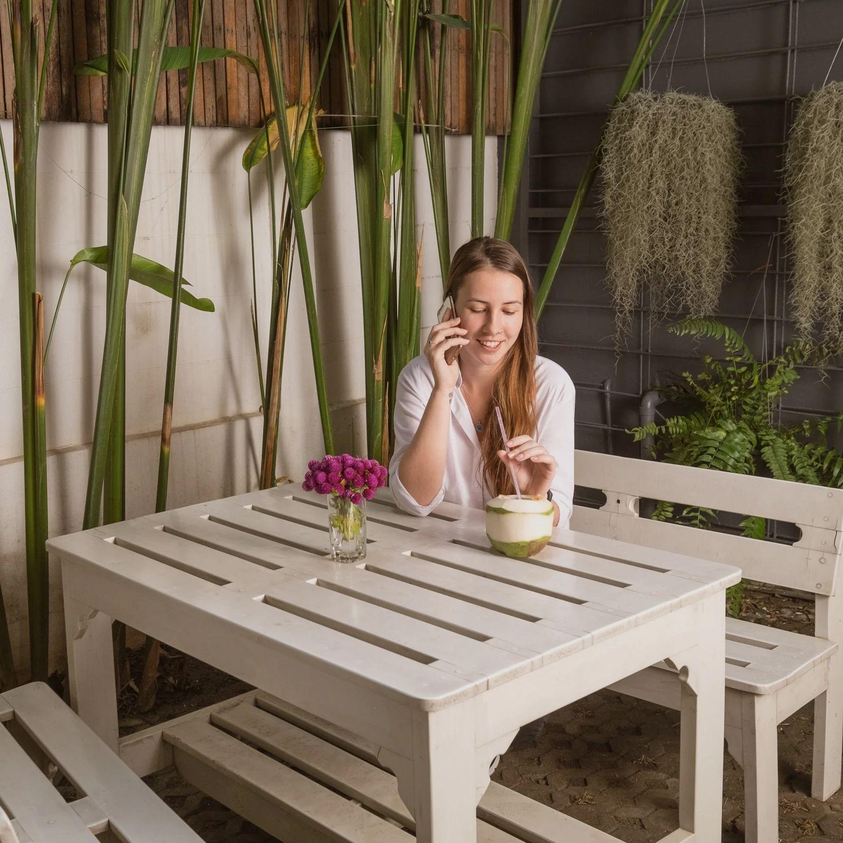 Woman sitting at a white outdoor table in the courtyard, talking on the phone with tropical plants in the background