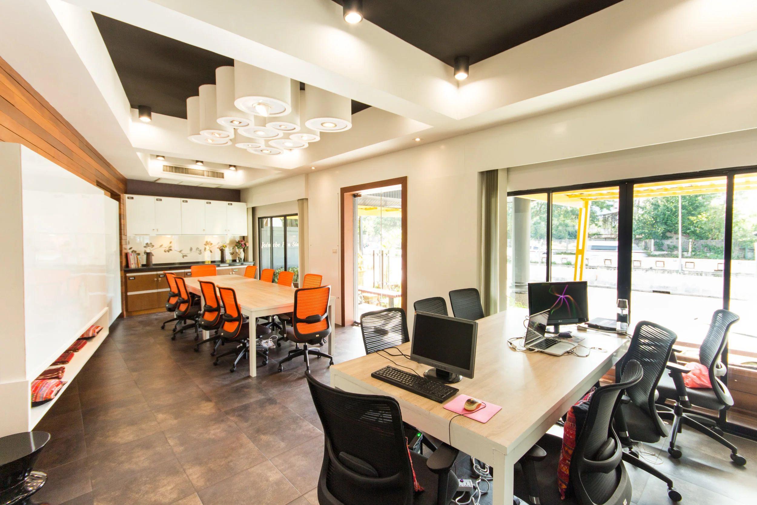 Open-plan coworking area with desks, computers, orange chairs and large windows overlooking the street