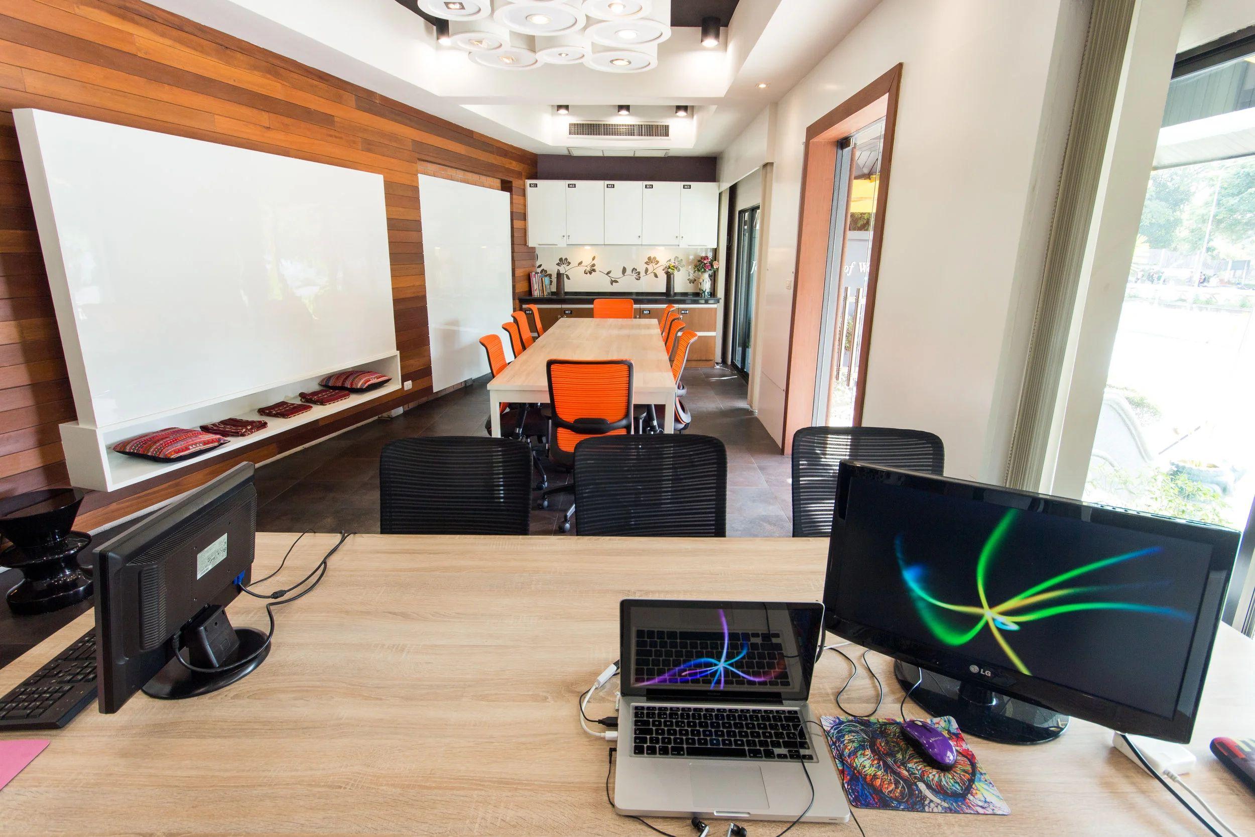 Coworking area with desks and computers in the foreground, conference room with orange chairs visible in the background
