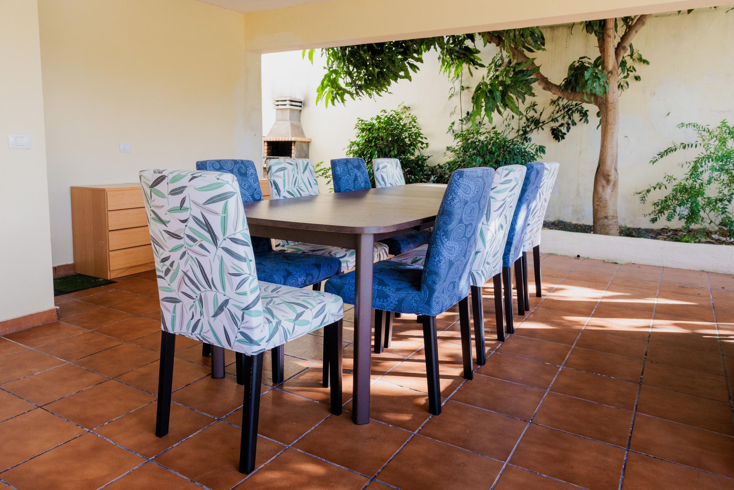 Covered outdoor dining terrace with a large table and colorful chairs surrounded by trees