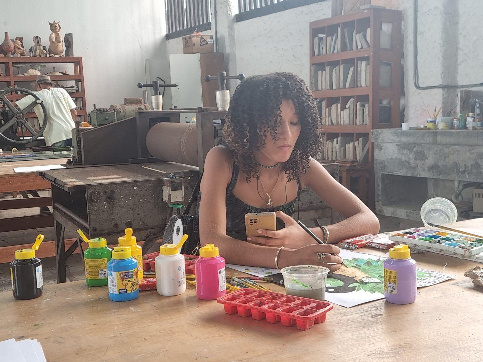 A person working on a painting at a table in an art workshop space, surrounded by art supplies and paint.