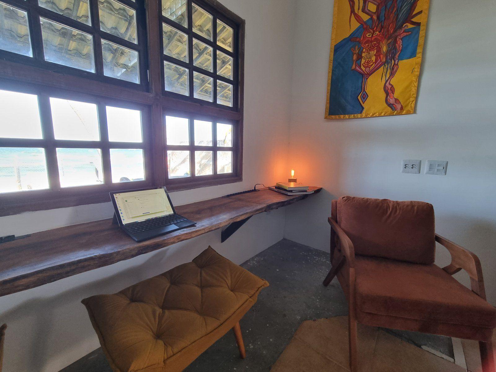 A wooden live-edge desk with a laptop and lamp, facing large windows with an ocean view, and an armchair beside it.