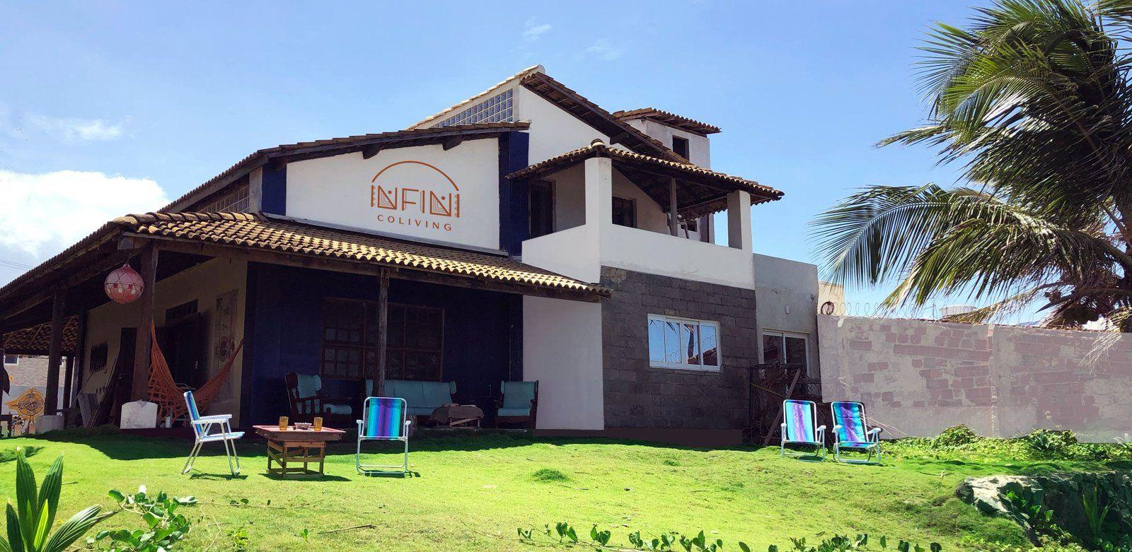 Exterior view of the Infini Coliving building with the Infini Coliving logo, green lawn, outdoor chairs, and ocean visible in the background.