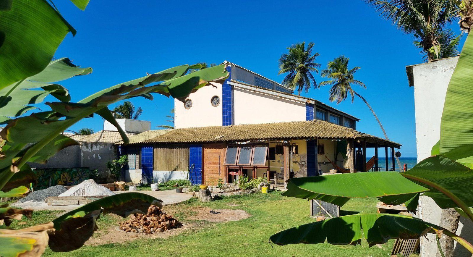 Exterior view of the Infini Coliving villa seen from the garden, with tropical plants, palm trees, and the ocean in the background.