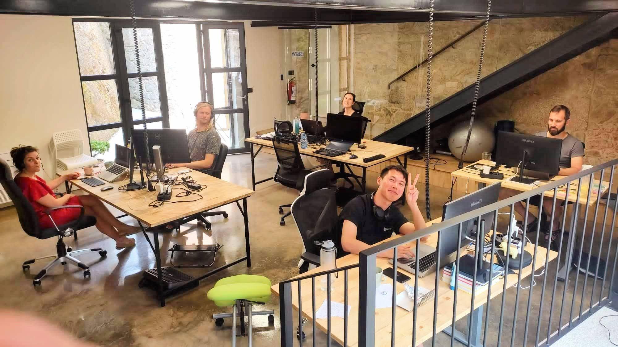 Multiple people working at desks in the coworking space with stone walls, monitors, and a staircase