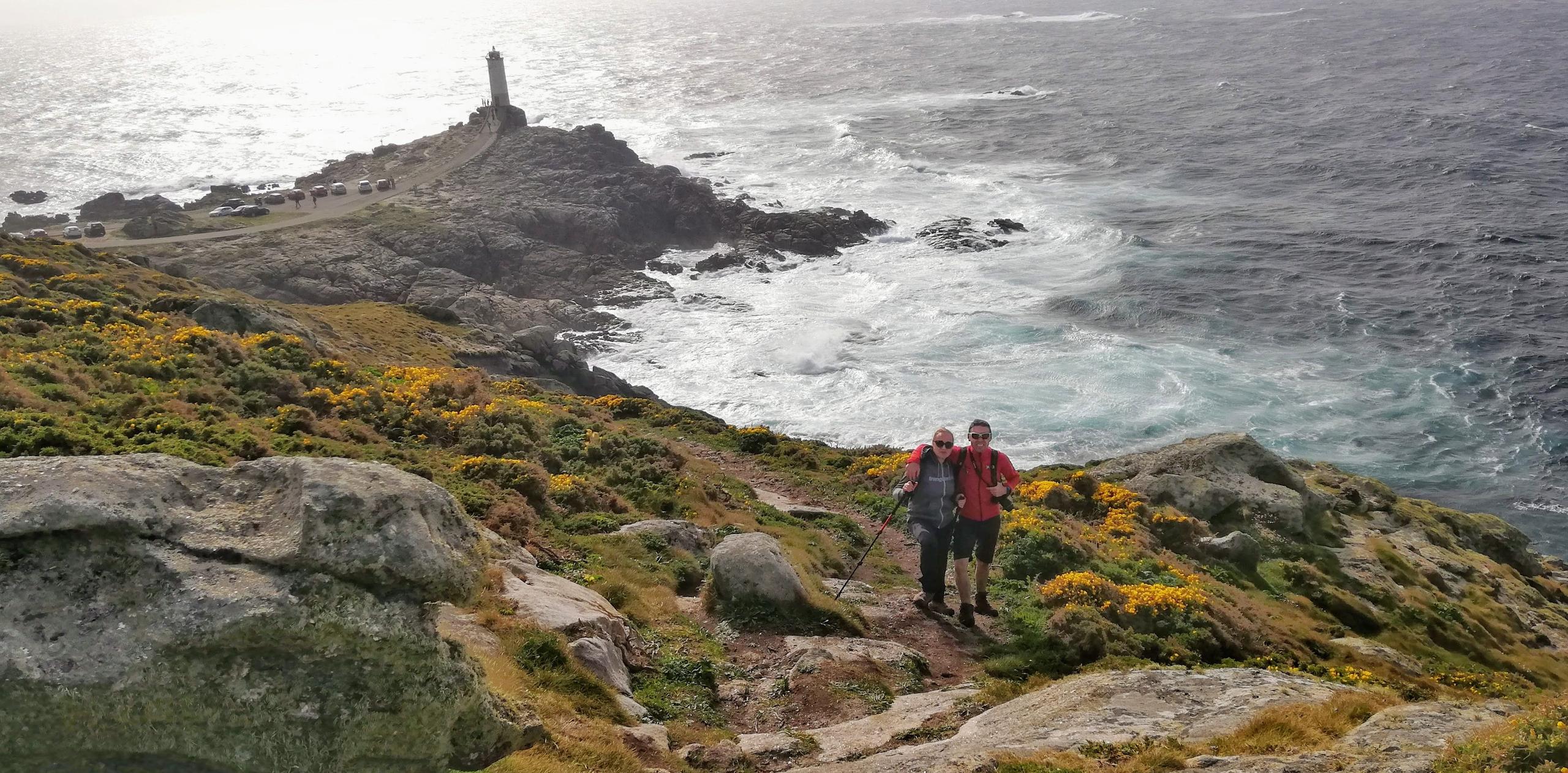 Two hikers on the Camiño dos Faros trail with Faro Roncudo lighthouse and the Atlantic Ocean in the background