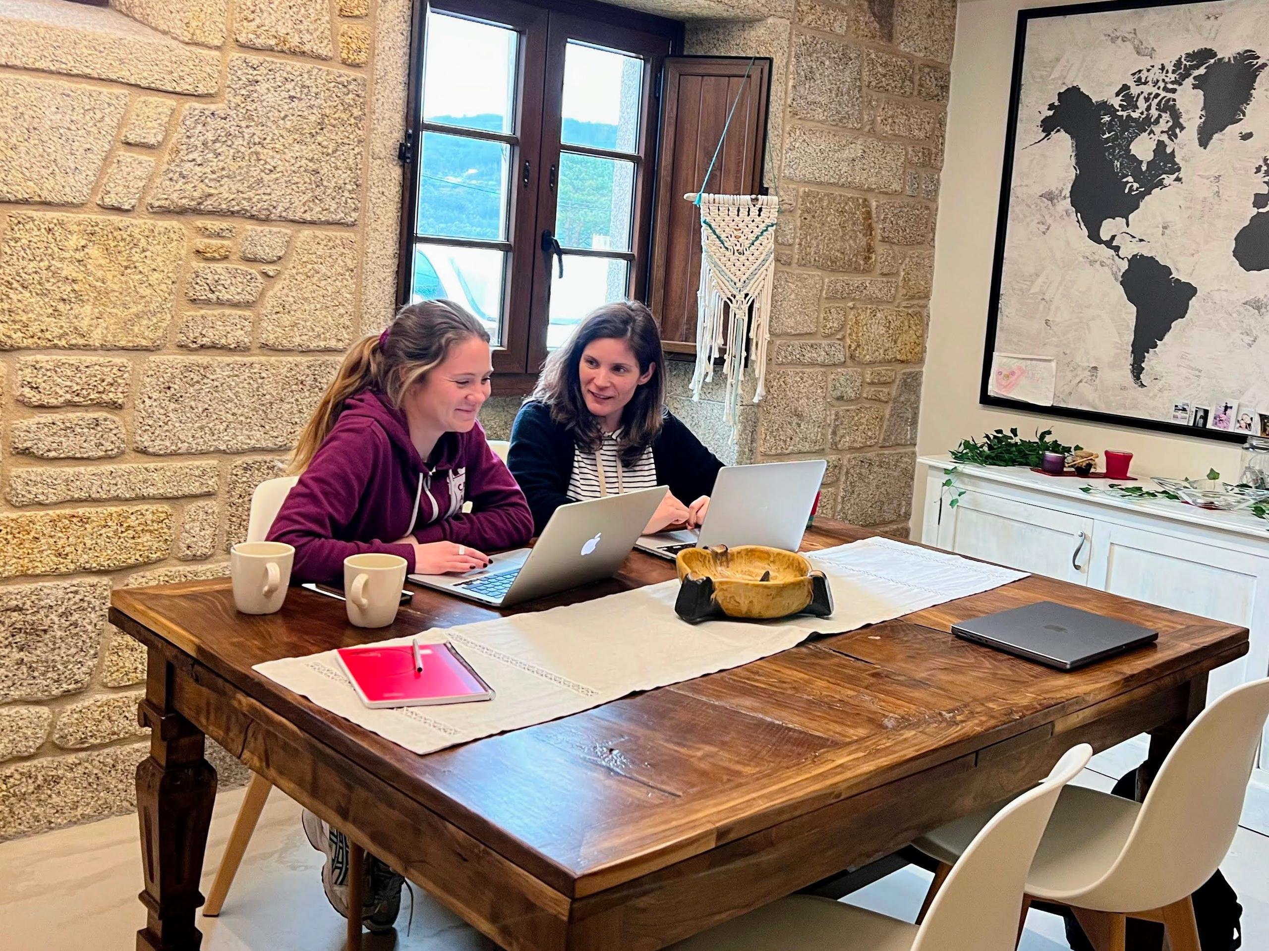 Two people working together on laptops at a wooden table in the coworking space with stone walls and a world map on the wall