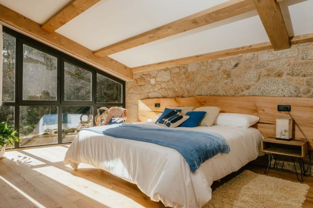 Family room O Cabanote with large double bed, wooden beams, stone wall, and large windows overlooking the garden