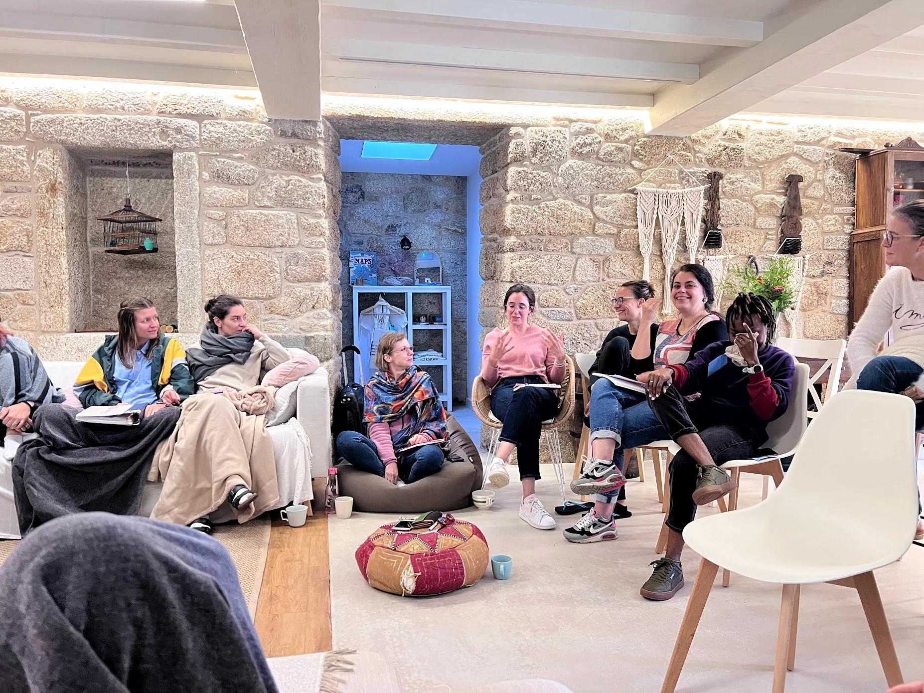 Group of people sitting in a circle in the living room for a community session or workshop, with stone walls and cosy decor