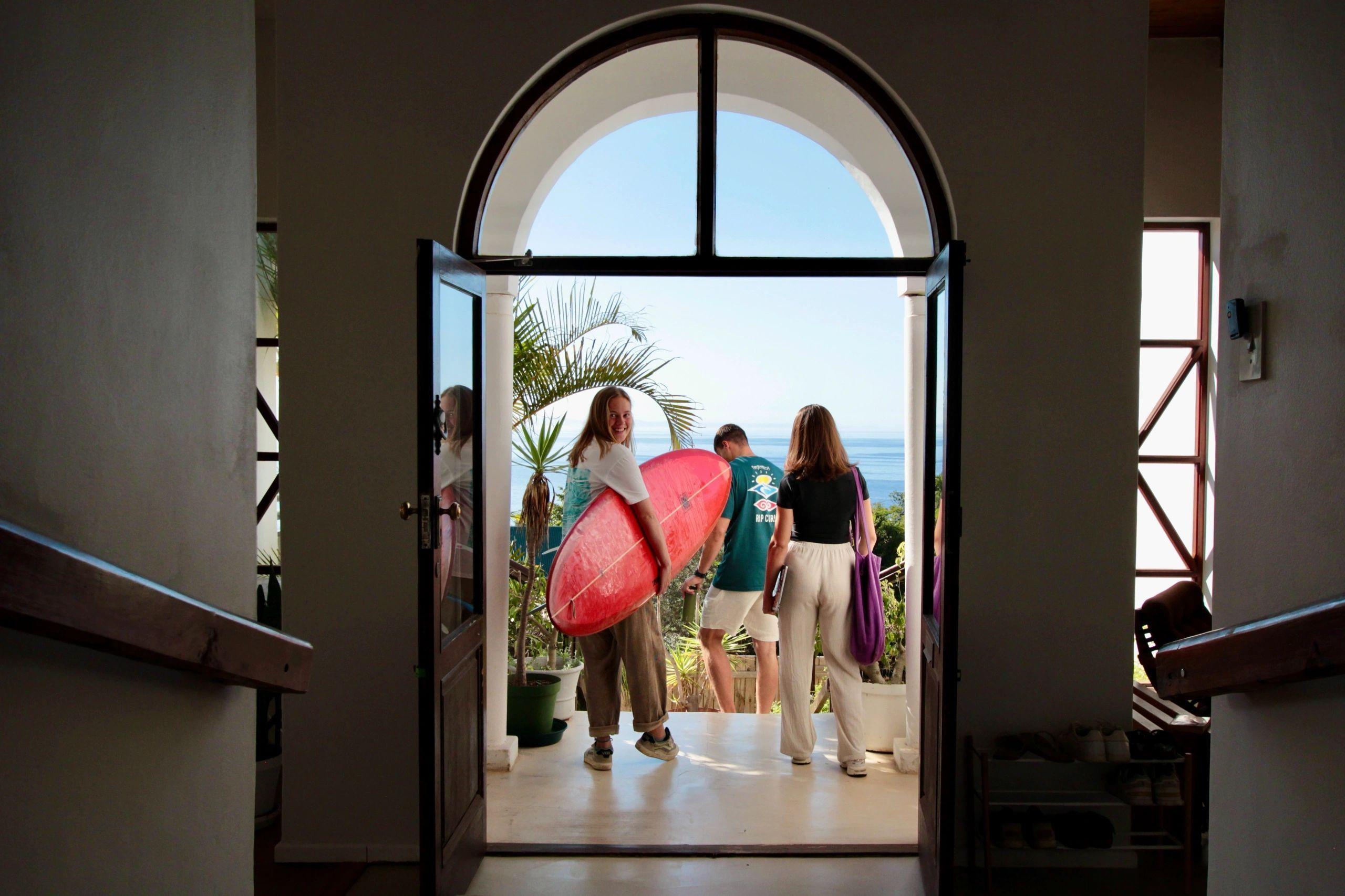 View from inside the house through an arched doorway, showing residents heading out with a surfboard, with the ocean visible in the background