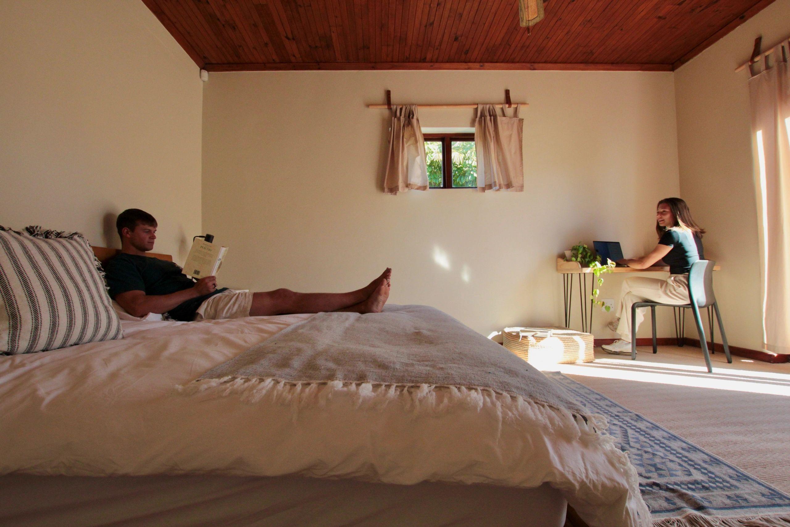 Couple in a bedroom with wooden ceiling — one reading on the bed, one working at a desk by the window