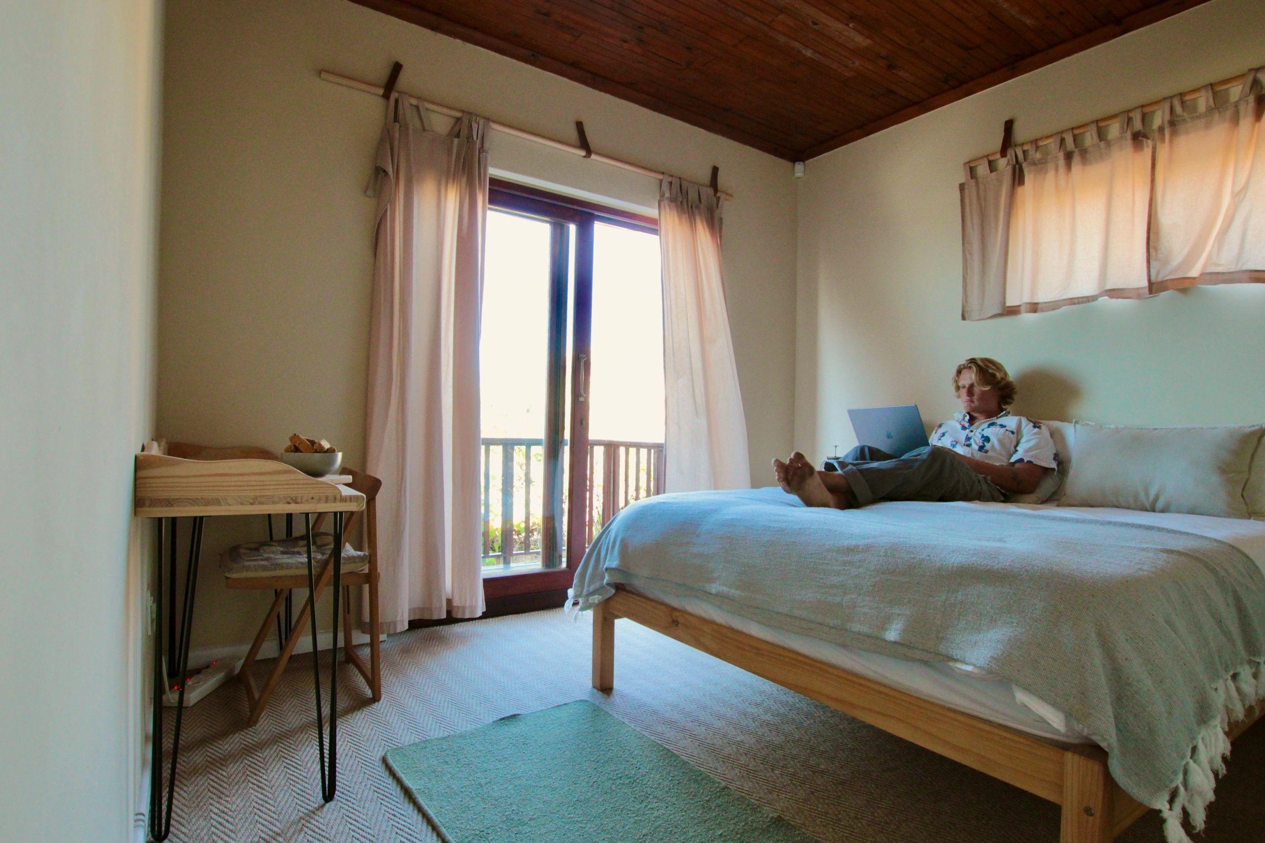 Resident lying on bed using a laptop in a room with wooden ceiling and balcony door