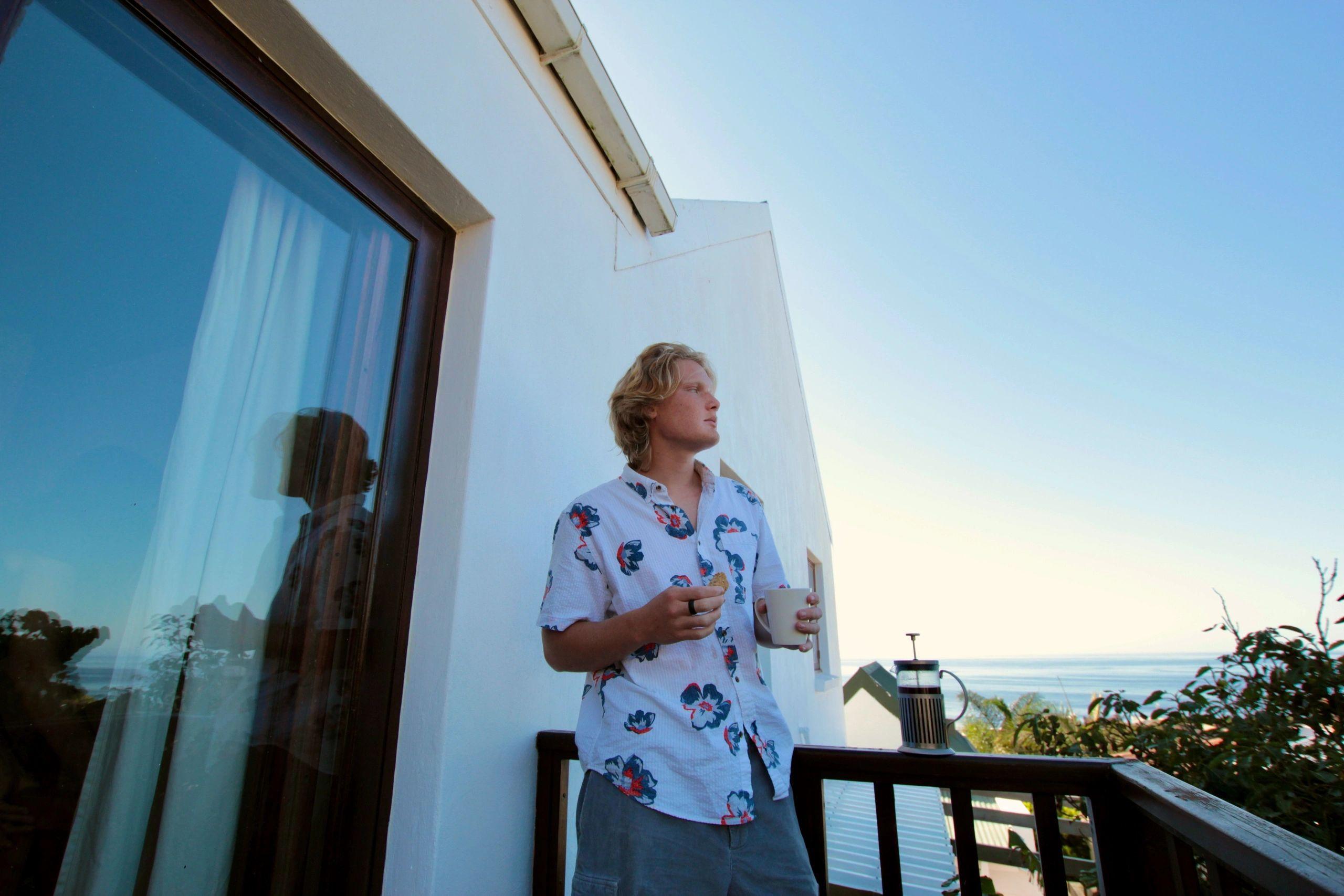 Resident standing on a private room terrace holding a coffee cup, looking out at the ocean view
