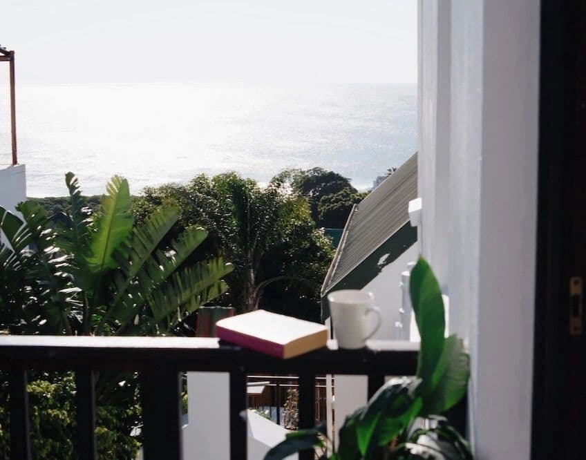 View from a private room terrace showing the ocean, tropical plants, and a book and coffee cup on the railing