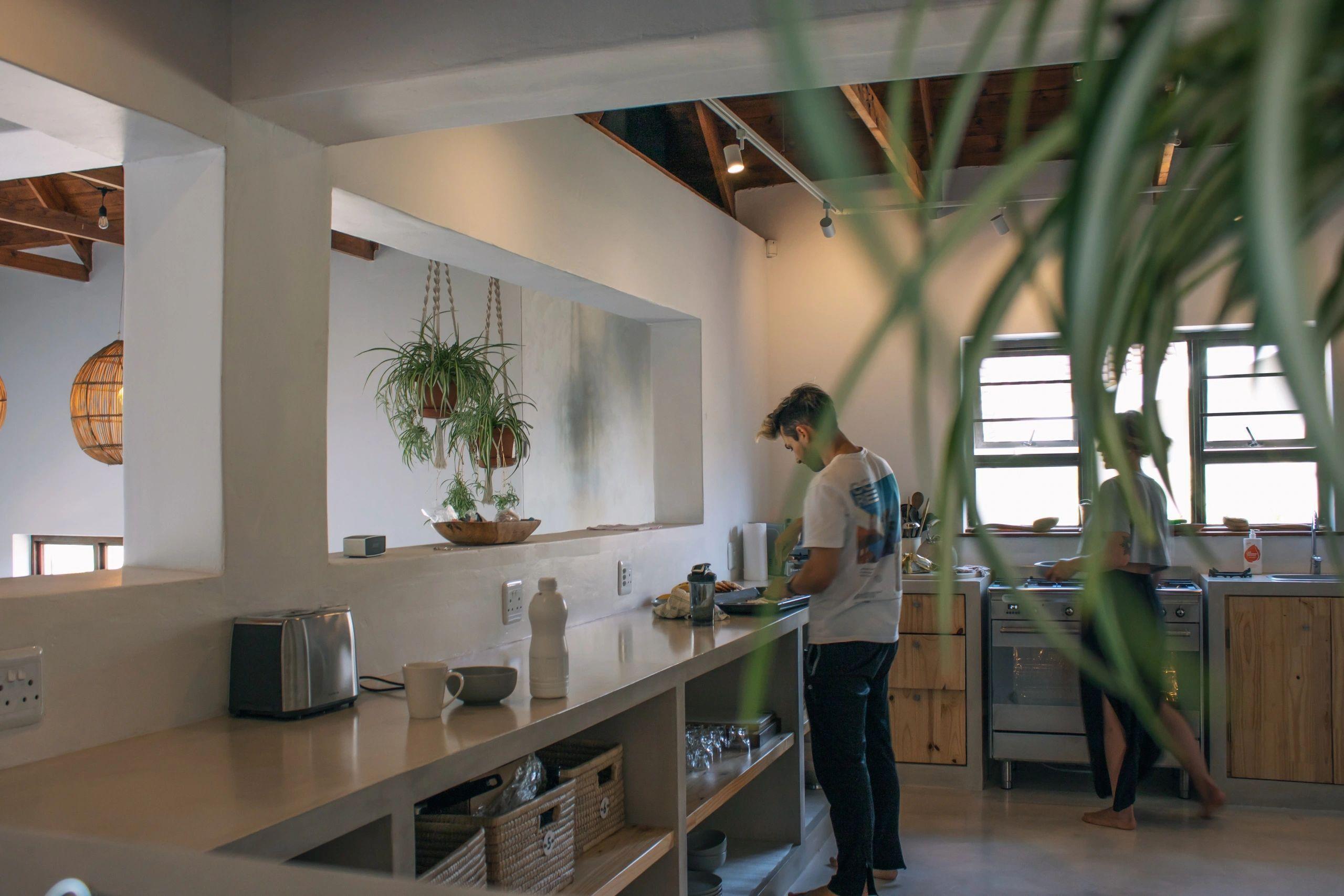 Resident preparing food in the communal kitchen, with long countertop, wicker baskets, hanging plants, and wooden beam ceiling visible