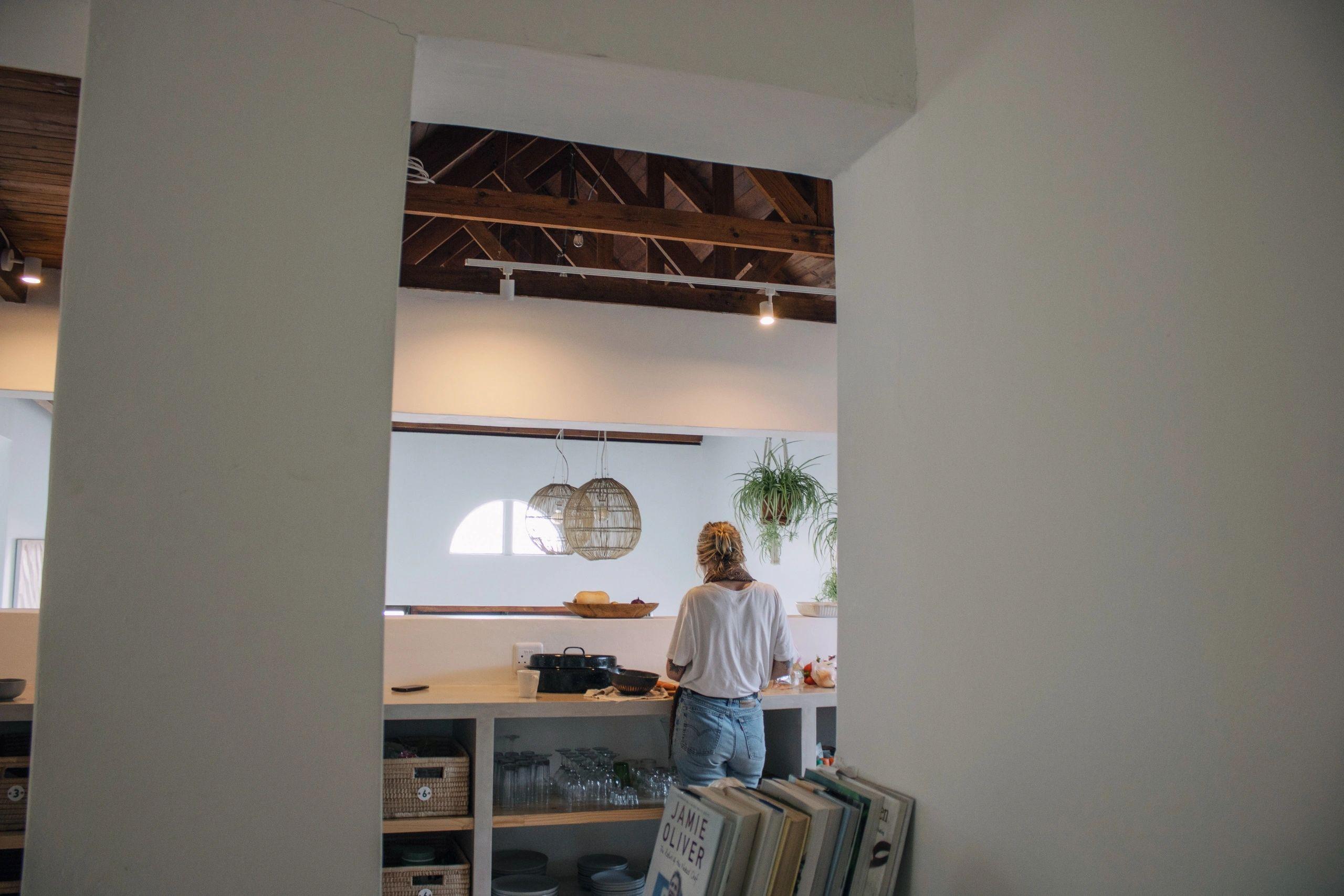 Person working in the communal kitchen, viewed through a doorway, with wooden beam ceiling, hanging pendant lights, and open shelving