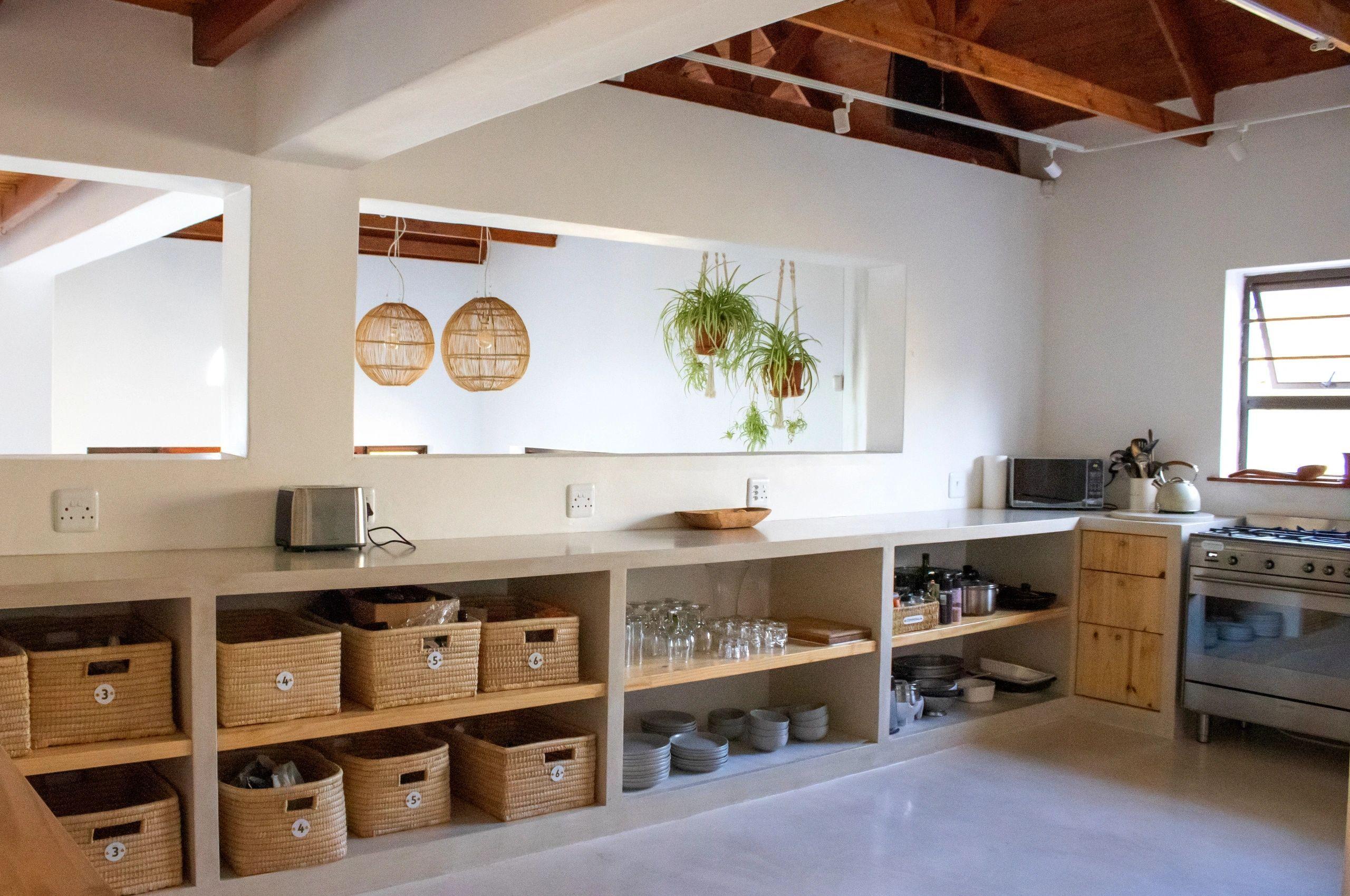 Wide view of the communal kitchen with long countertop, wicker storage baskets, hanging pendant lights, plants, and cooking appliances