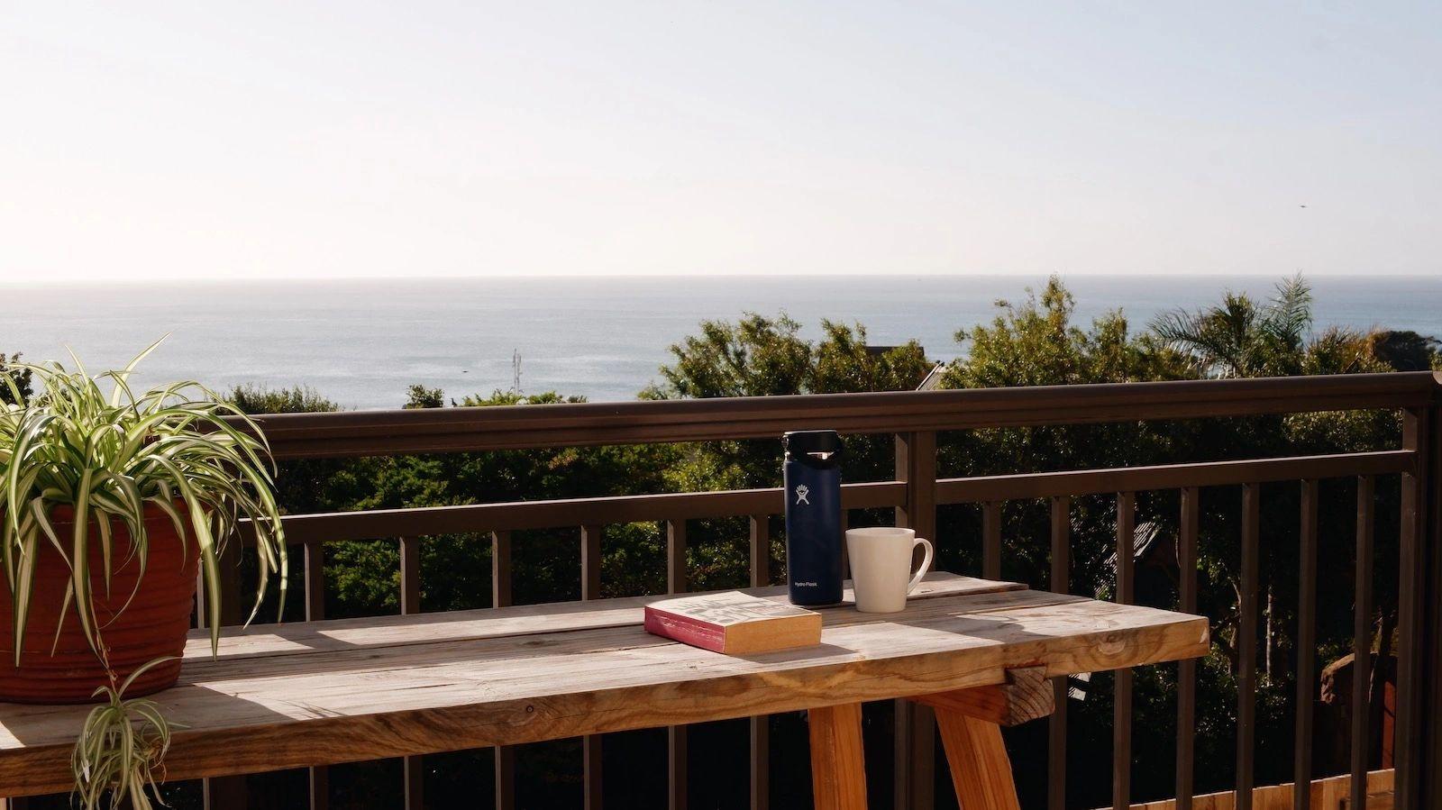 Wooden outdoor deck with a table, book, and water bottle, overlooking the ocean through lush greenery