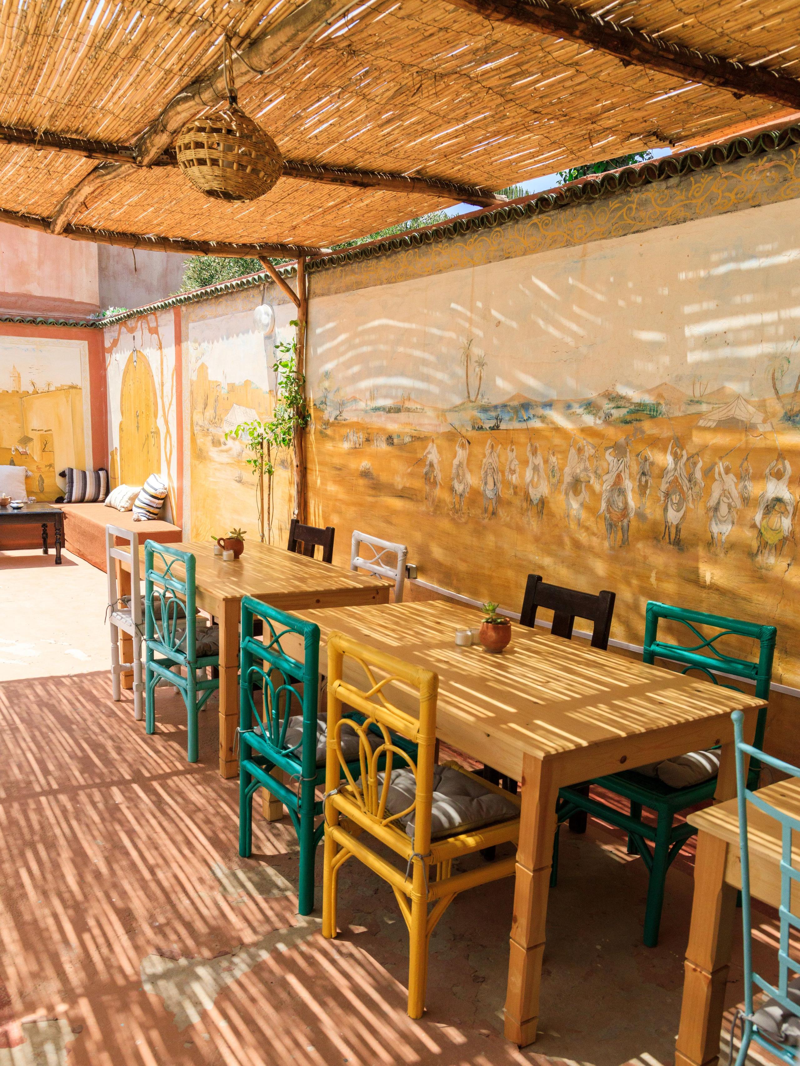 Outdoor dining area with wooden tables and colorful chairs under a bamboo canopy with Moroccan mural