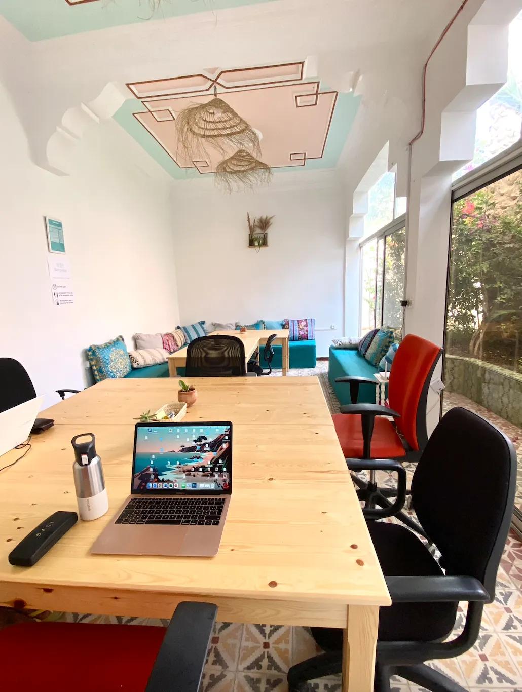 Indoor coworking room with wooden desks, office chairs, Moroccan sofas, and large windows overlooking the garden