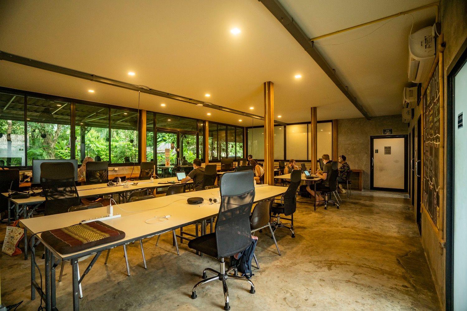Large air-conditioned indoor coworking room with long desks, ergonomic chairs, and floor-to-ceiling windows overlooking the tropical garden