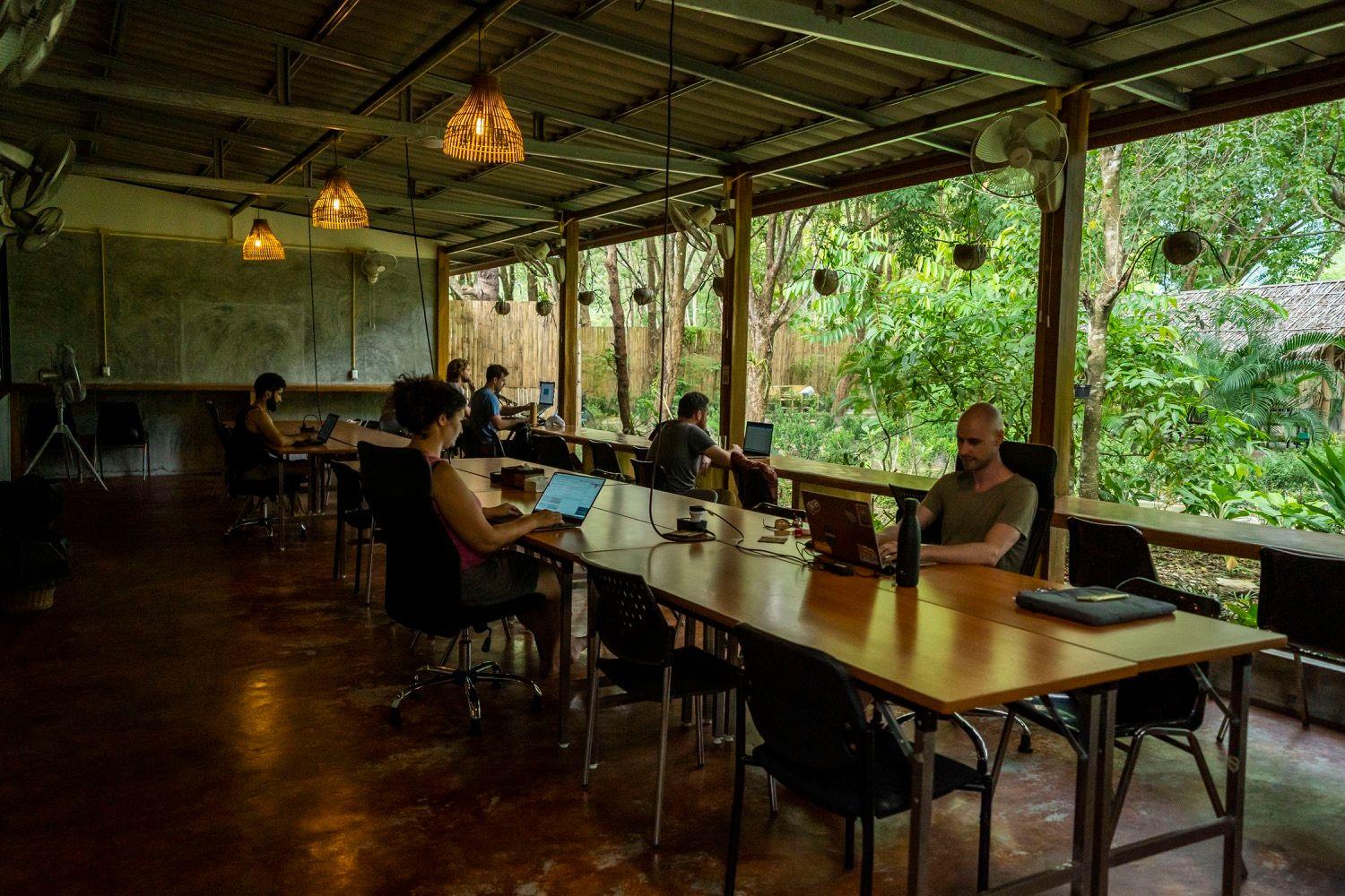 Open-air coworking room with long wooden desks, pendant lights, and views of the tropical garden