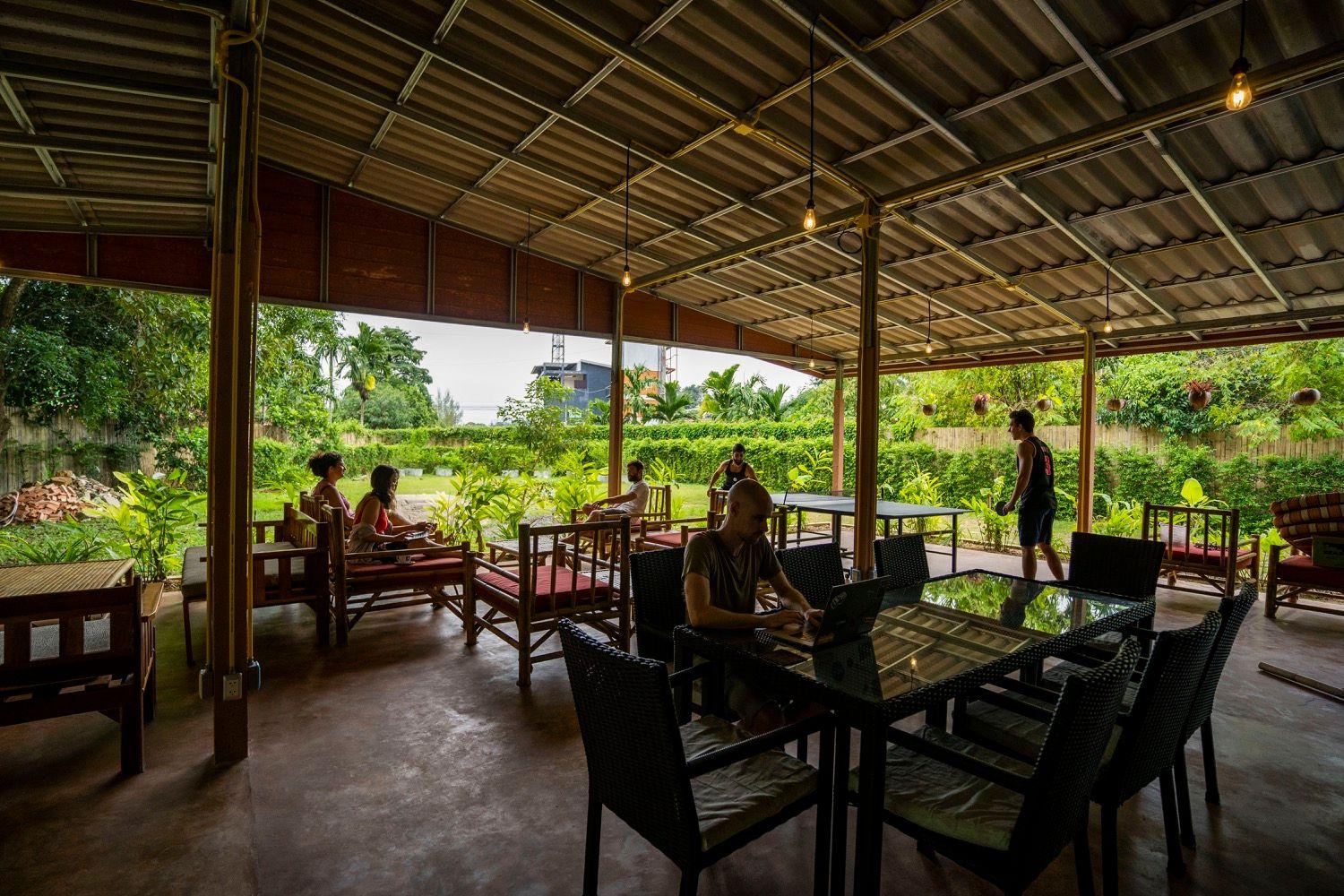 Open-air communal dining area with members sitting at tables surrounded by tropical garden
