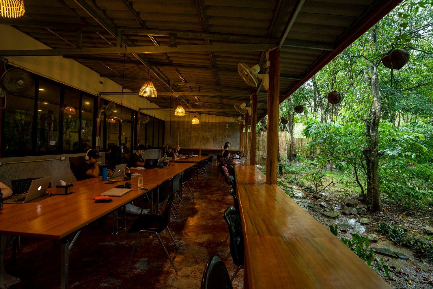 Long open-air coworking deck with wooden desks, pendant lights, and tropical garden views
